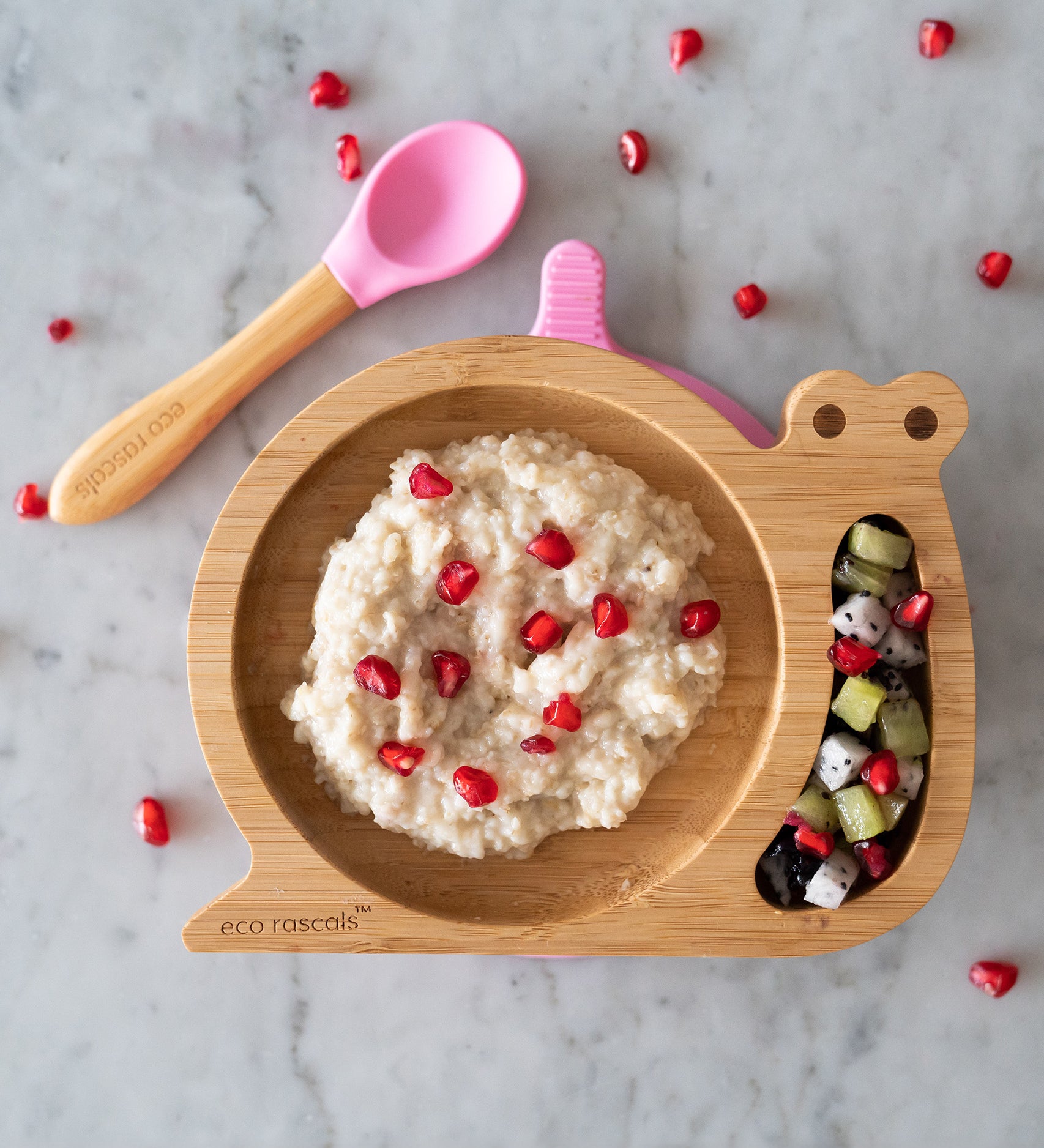 Eco Rascals Snail Bamboo Suction Plate with pink suction base, filled with rice pudding and a fruit salad with pomegranate seeds and a pink silicone and bamboo baby spoon. 