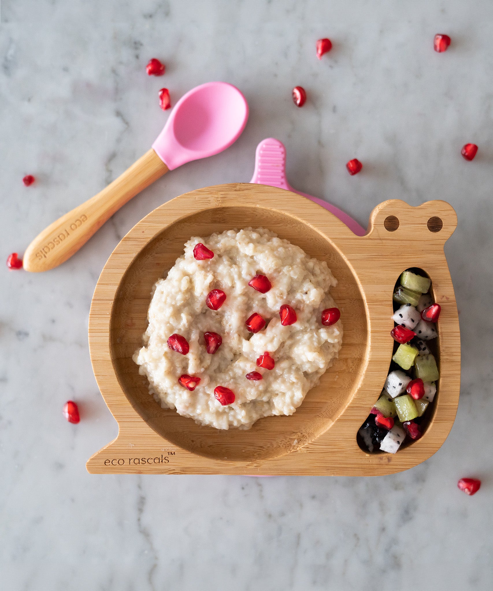 Eco Rascals Snail Bamboo Suction Plate with pink suction base, filled with rice pudding and a fruit salad with pomegranate seeds and a pink silicone and bamboo baby spoon. 