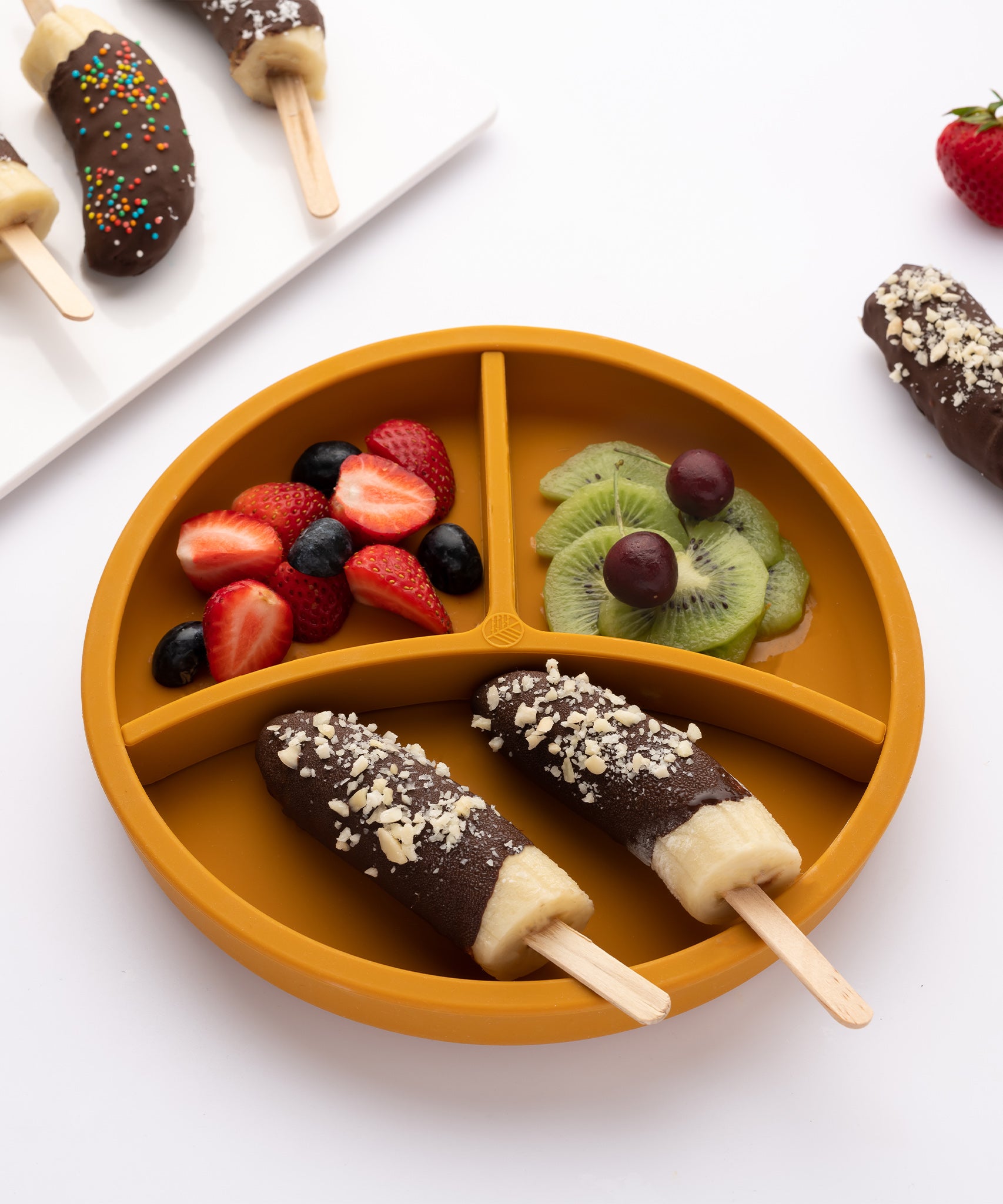 Eco Rascals Silicone Baby Plate in mustard with removable divider, with banana lollies and fresh fruit and berries. 