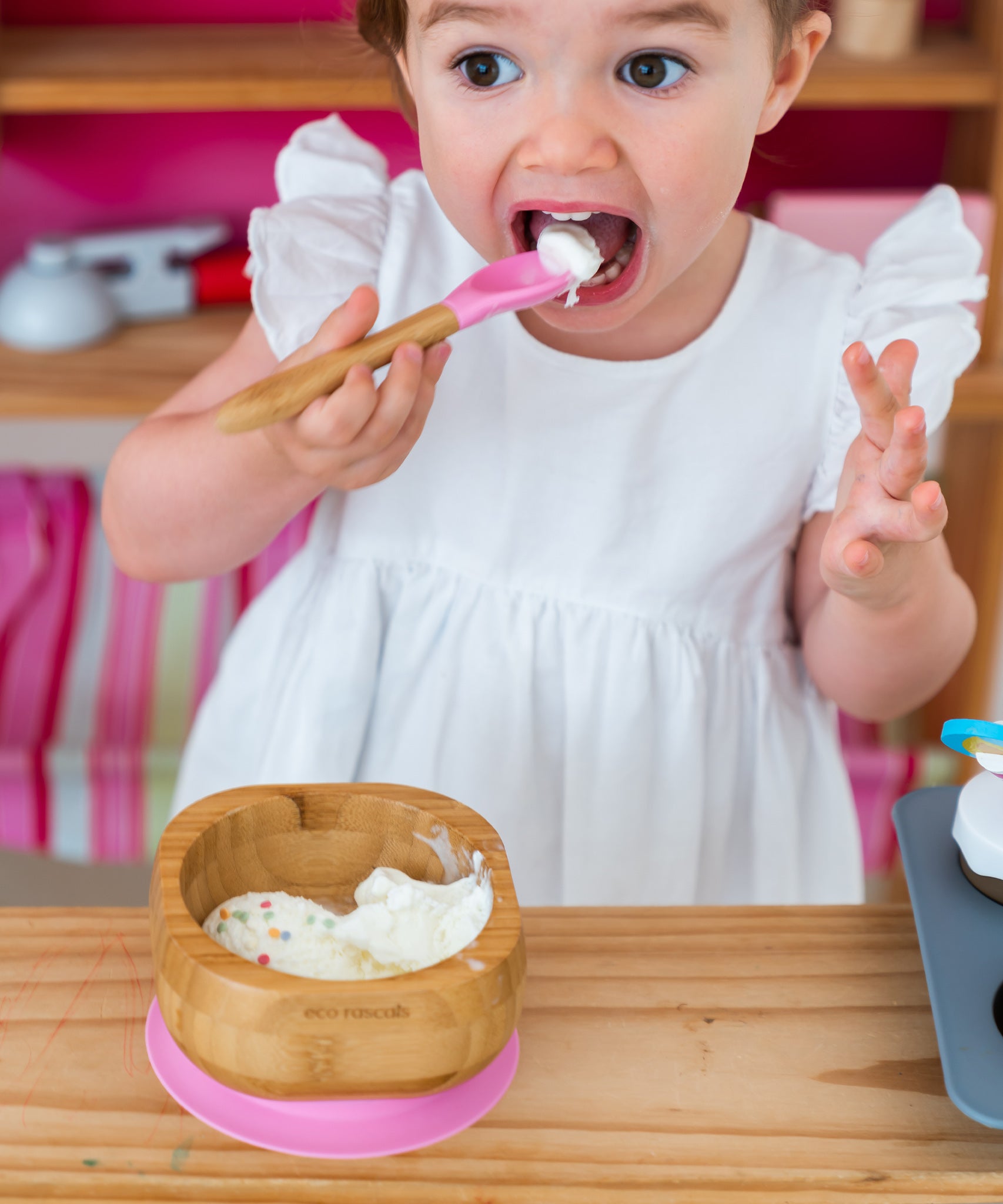 Toddler wearing a white dress eating icecream at a wooden table from the Eco Rascals Bamboo Bowl & Spoon Weaning Set in pink silicone.