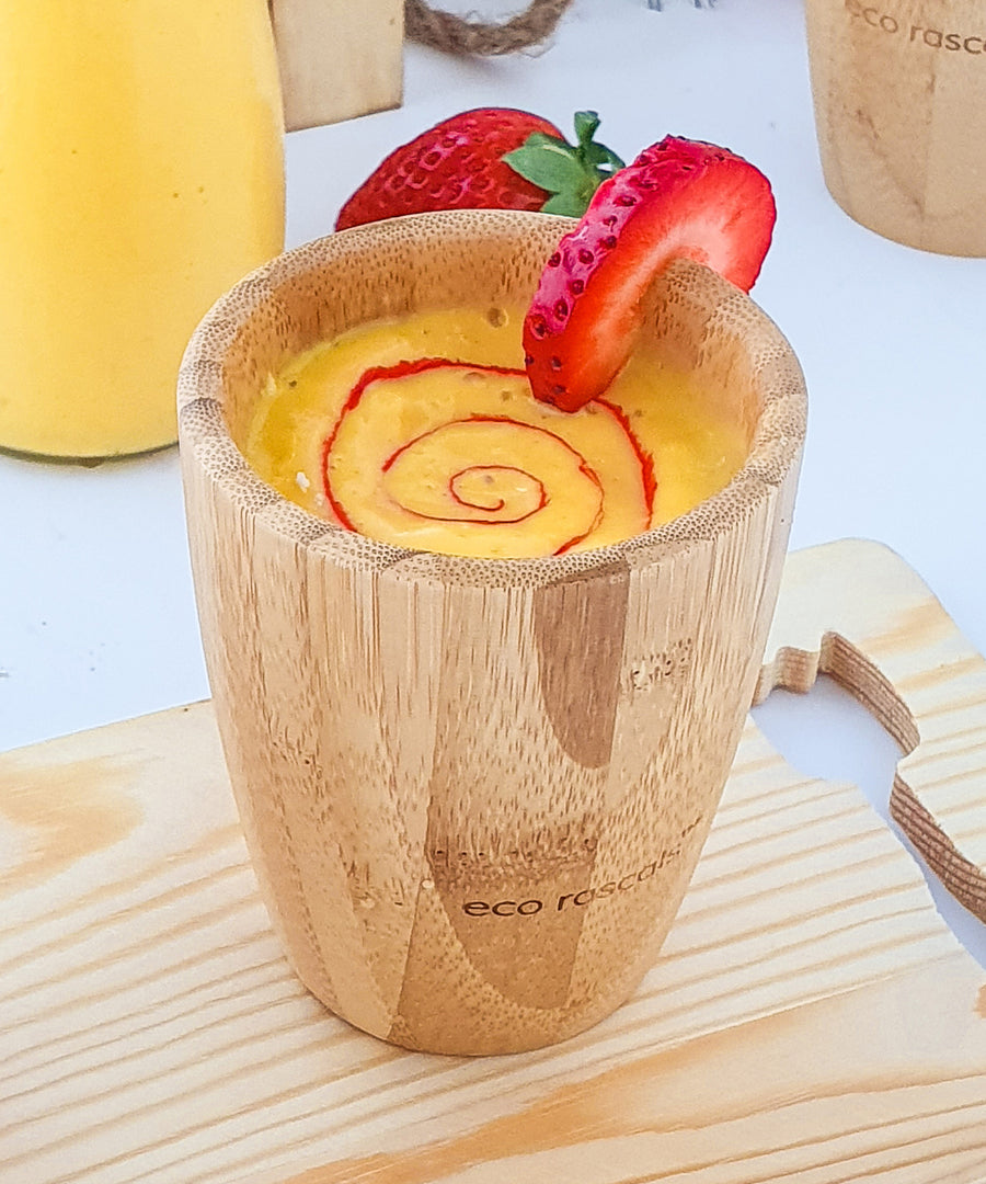 A yellow mango smoothie with red strawberry puree swirl in a bamboo baby cup, decorated with strawberry slices..