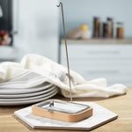 The Ecoliving Dish Brush holder with soap try is stood on a hexagonal tile, which is set on a wooden table. In the background are 4 plates stacked on top of each other with a dish cloth on top