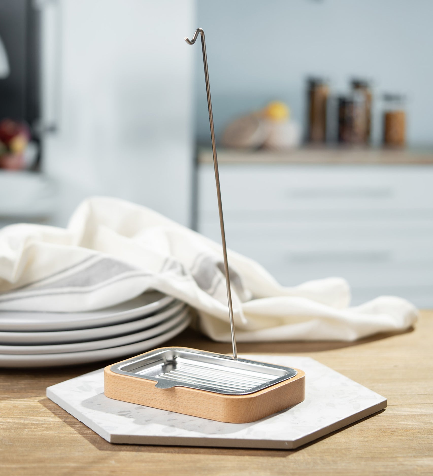 The Ecoliving Dish Brush holder with soap try is stood on a hexagonal tile, which is set on a wooden table. In the background are 4 plates stacked on top of each other with a dish cloth on top