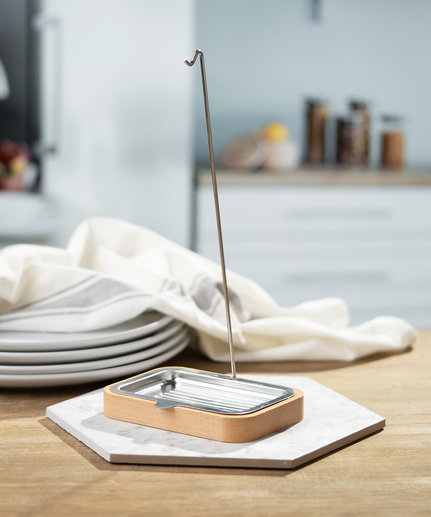 The Ecoliving Dish Brush holder with soap try is stood on a hexagonal tile, which is set on a wooden table. In the background are 4 plates stacked on top of each other with a dish cloth on top