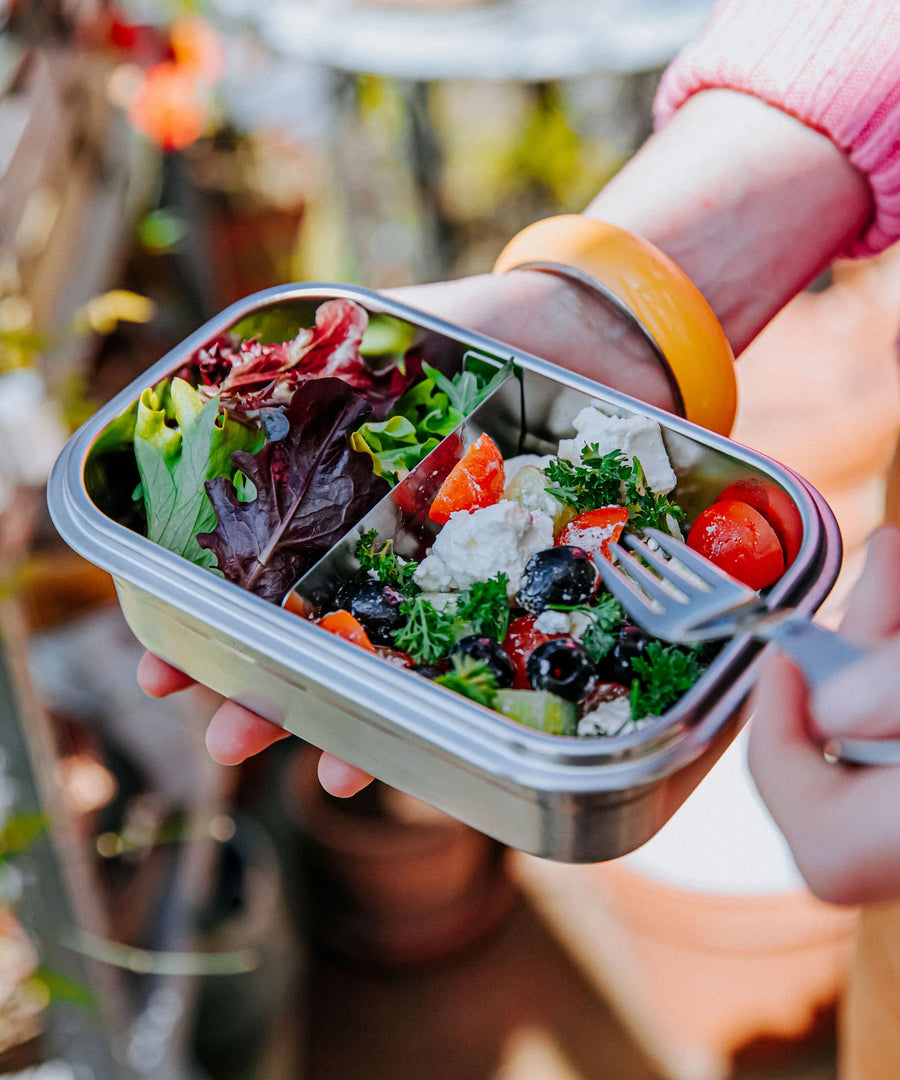 A close up of the Elephant Box Clip & Seal Lunchbox With Removable Divider - 800ml with a salad inside. A person is holding the lunch box in one hand, and a Elephant Box Spork in the other