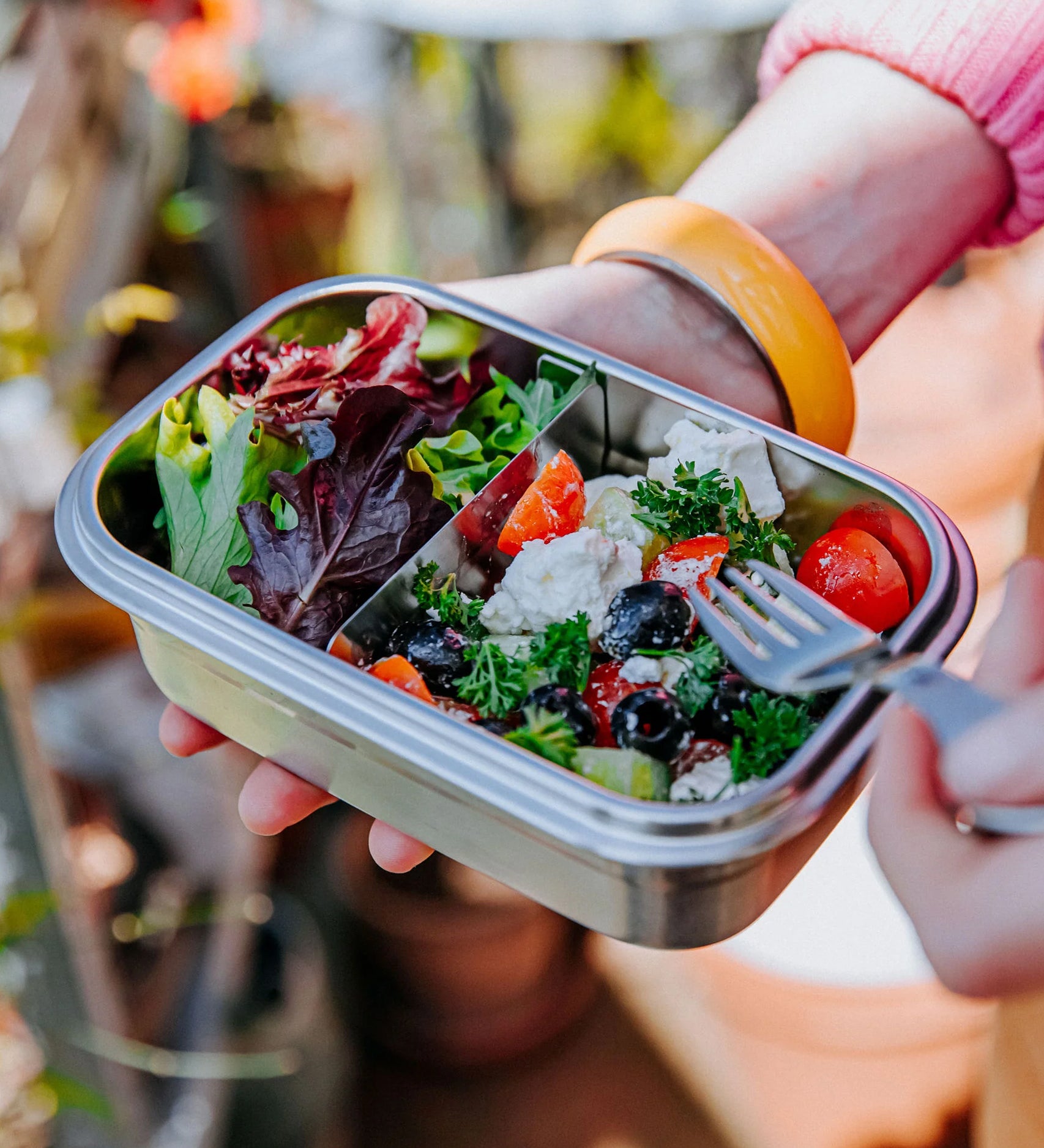 A close up of the Elephant Box Clip & Seal Lunchbox With Removable Divider - 800ml with a salad inside. A person is holding the lunch box in one hand, and a Elephant Box Spork in the other