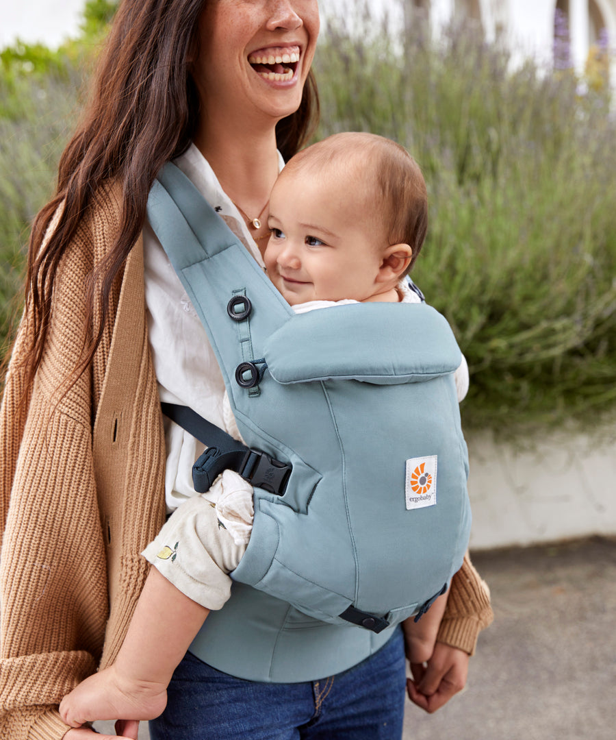 Woman with long brown hair, laughing, standing in a courtyard wearing her baby in a Ergobaby Adapt  SoftTouch™  Cotton Baby Carrier in Slate Blue.