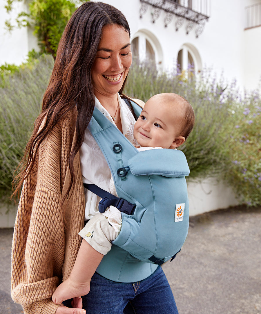 Woman with long brown hair standing in a courtyard wearing her baby in a Ergobaby Adapt  SoftTouch™  Cotton Baby Carrier in Slate Blue.