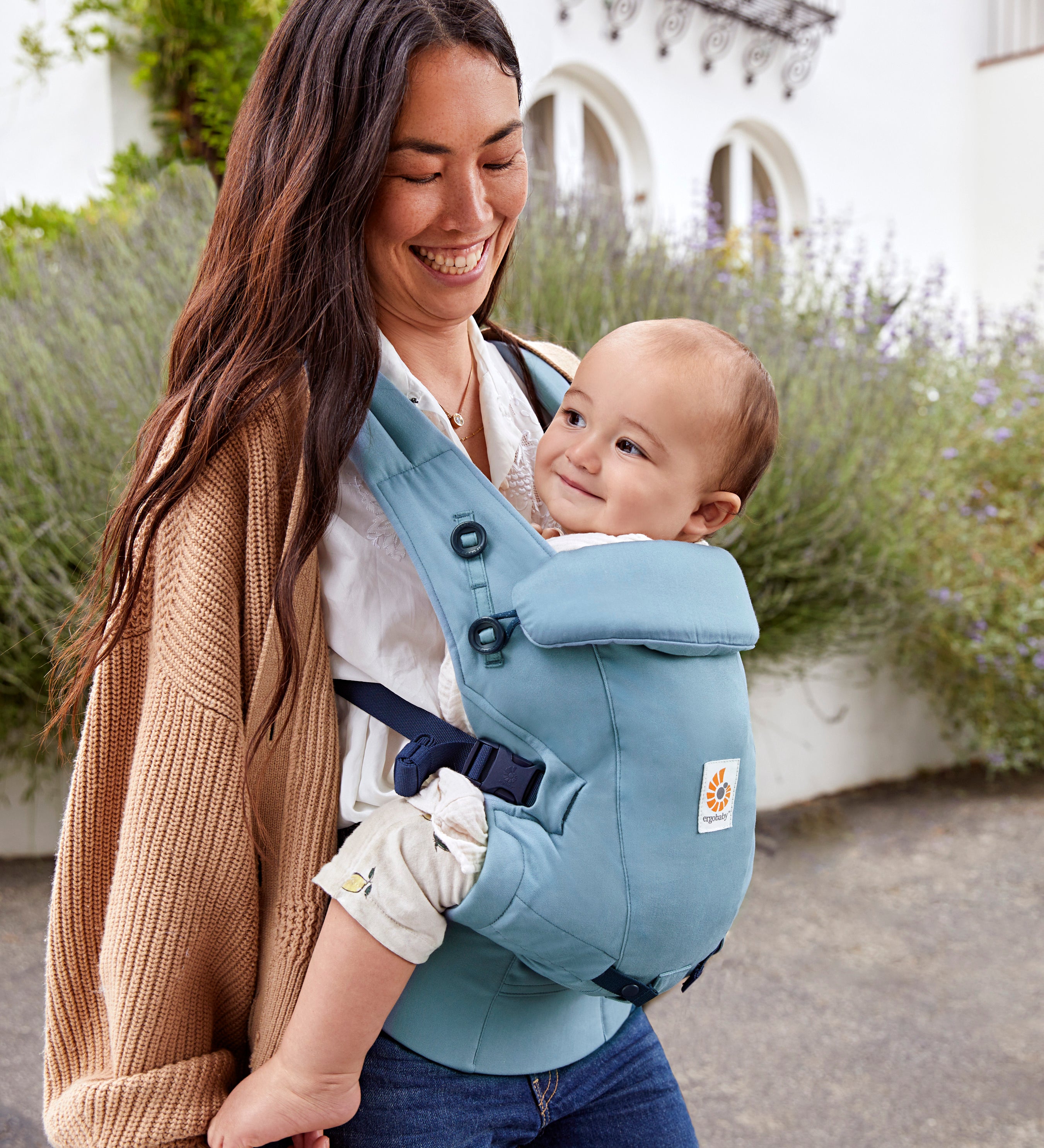 Woman with long brown hair standing in a courtyard wearing her baby in a Ergobaby Adapt  SoftTouch™  Cotton Baby Carrier in Slate Blue.