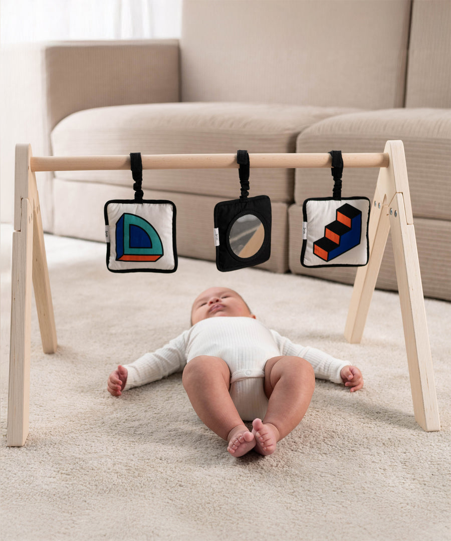 A baby lying down on their back, underneath a wooden baby gym, with the Etta Loves x Camille Walala Sensory Hanging Squares attached to the rung across the top of the baby gym. The baby is looking upwards towards the sensory squares, looking at the shapes and prints