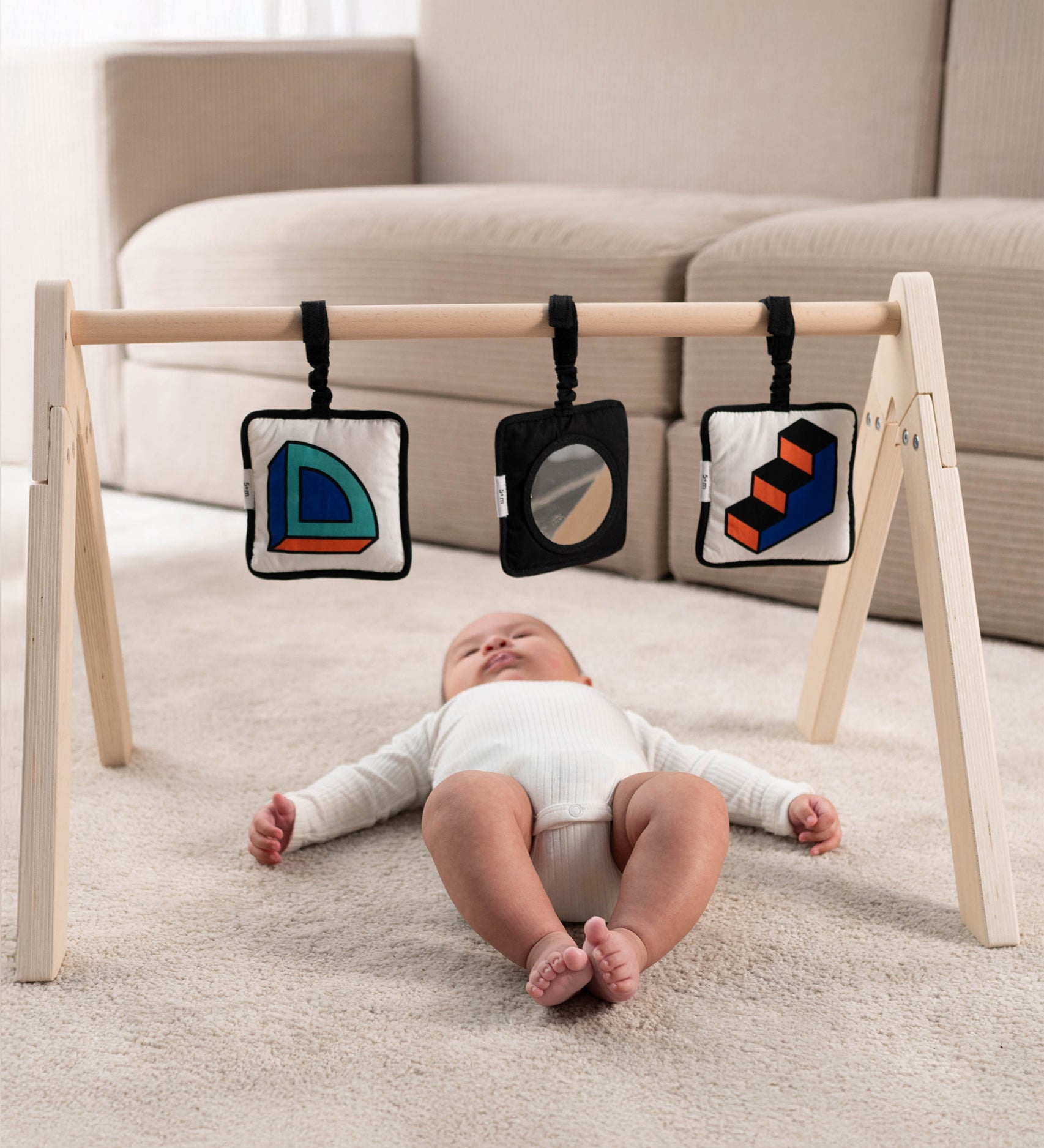 A baby lying down on their back, underneath a wooden baby gym, with the Etta Loves x Camille Walala Sensory Hanging Squares attached to the rung across the top of the baby gym. The baby is looking upwards towards the sensory squares, looking at the shapes and prints