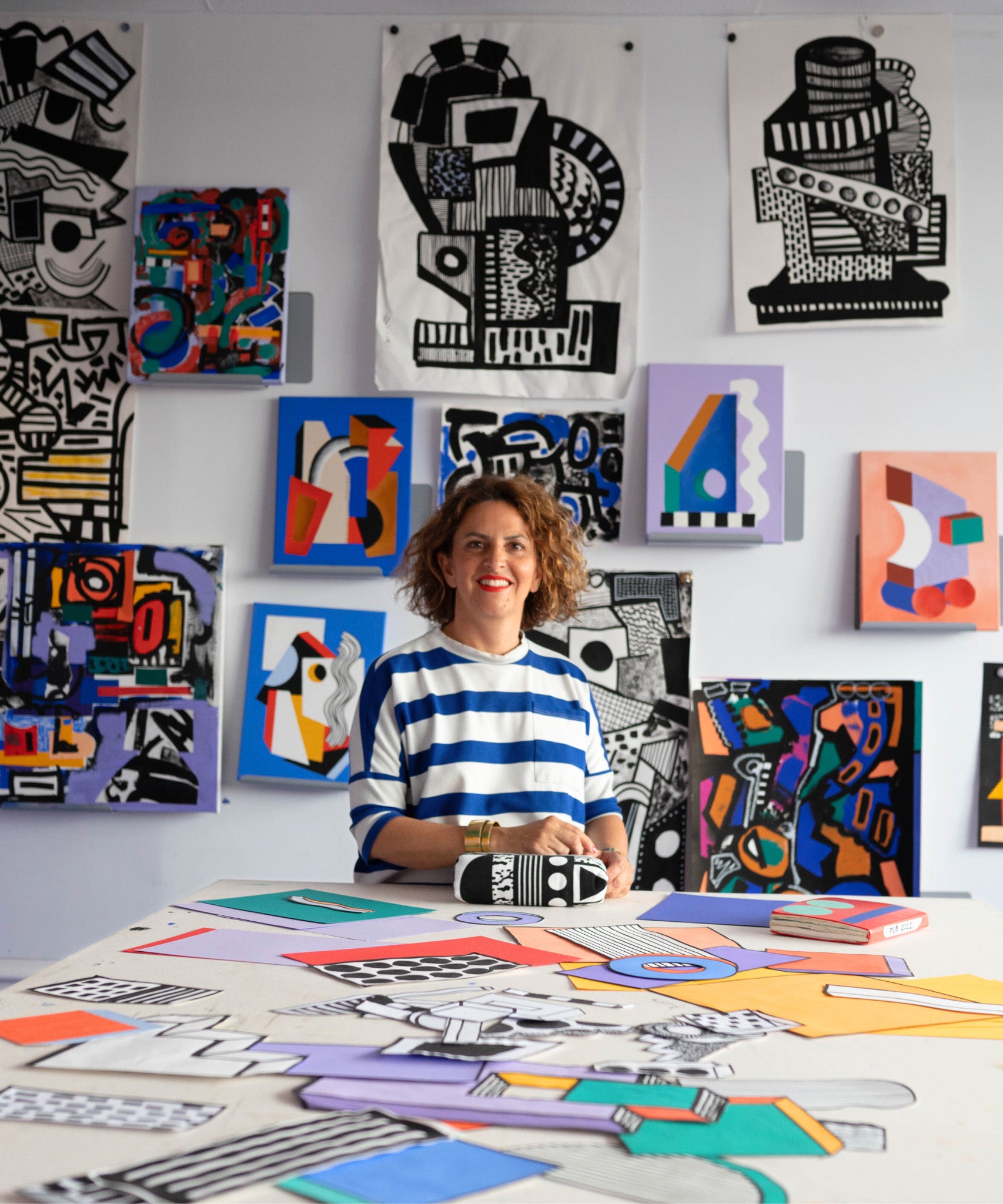 The artist Camille Walala surrounded by their artwork on a large table and a tall wall behind them. The artwork consists of colourful shapes and patterns, and monochromatic, detailed patterns