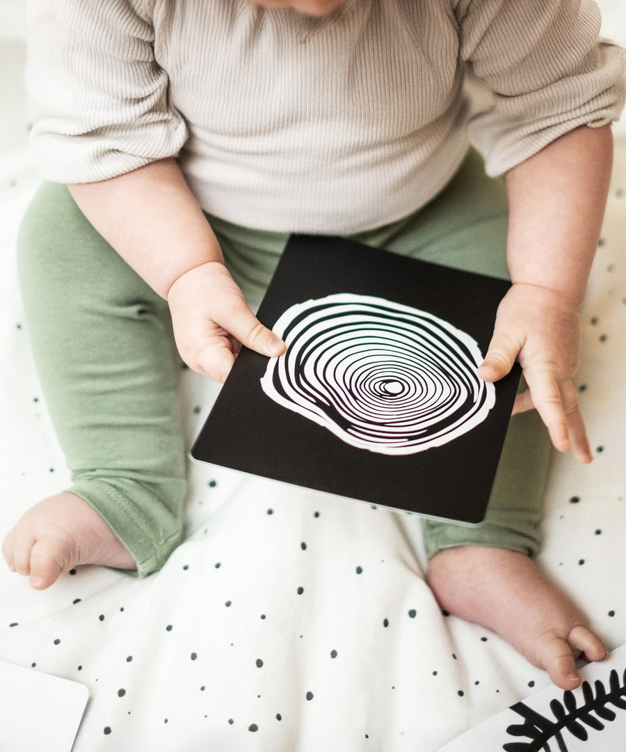 A baby playing with the Etta Loves Sensory Positive Affirmation Flash Cards. The card being held shows a black and white spiral 