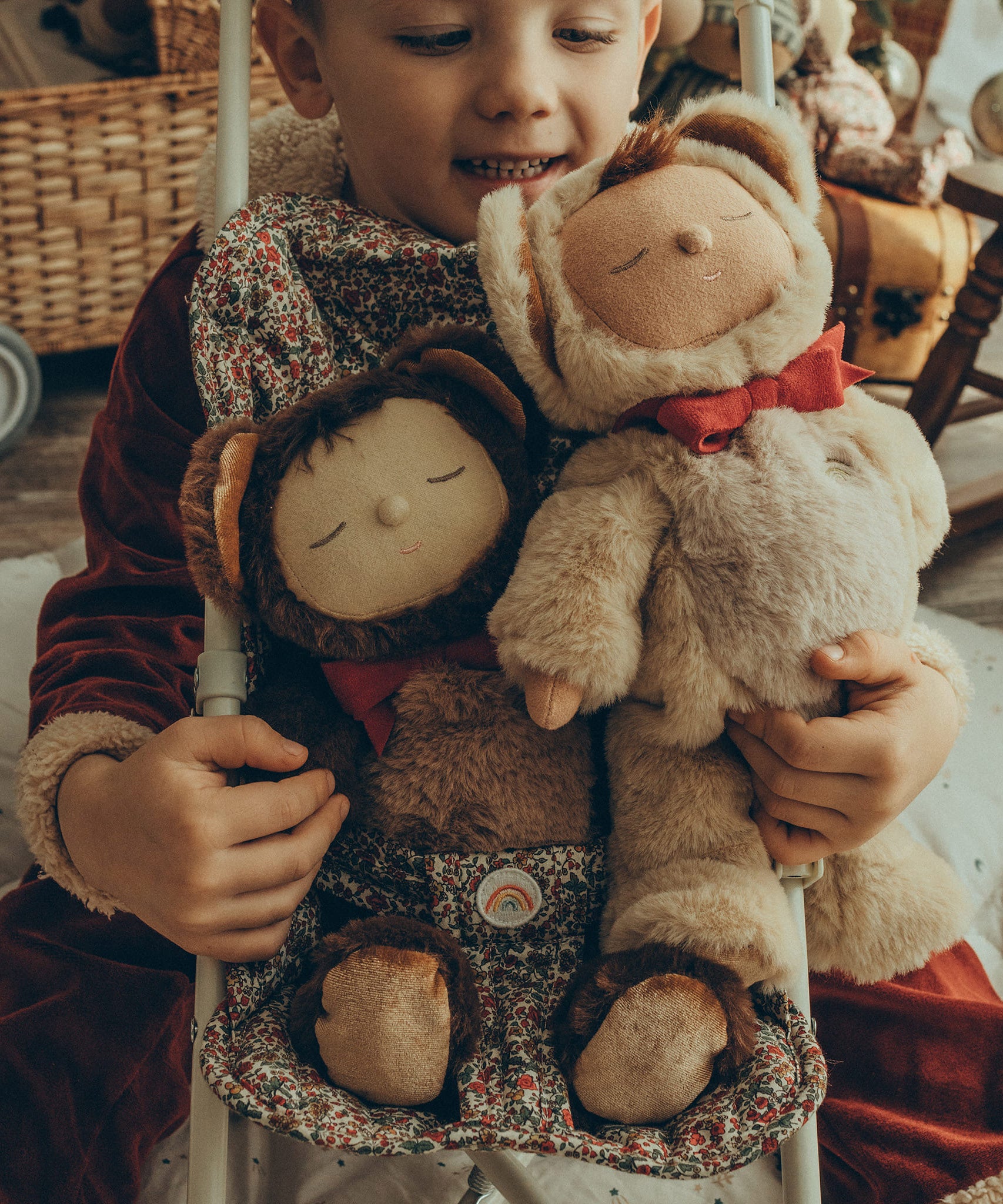 Olli Ella Cozy Dinkum Dolls Teddy Twiggy and Teddy Crumble together, sat in an Olli Ella Stroller with a child sat behind holding onto the back of the stroller. The child is wearing a red Santa outfit and is smiling down at Teddy Crumble