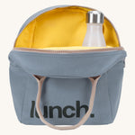 Fluf zip lunch bag in a light blue colour with a brown handle and a yellow inner lining holding a bottle on acream background.