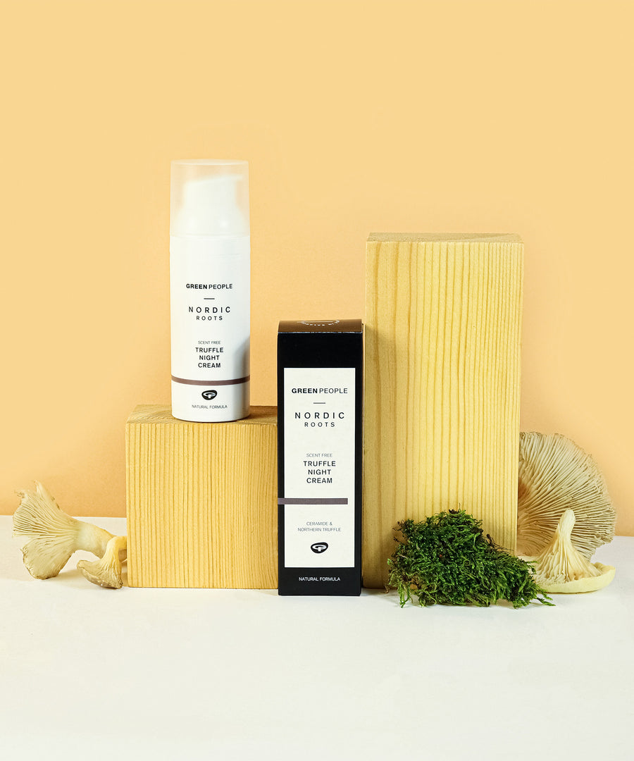 The Green People Nordic Roots Truffle Night Cream and its box is on a yellow background with the cream tube on a wooden block, with moss and mushrooms surrounding 