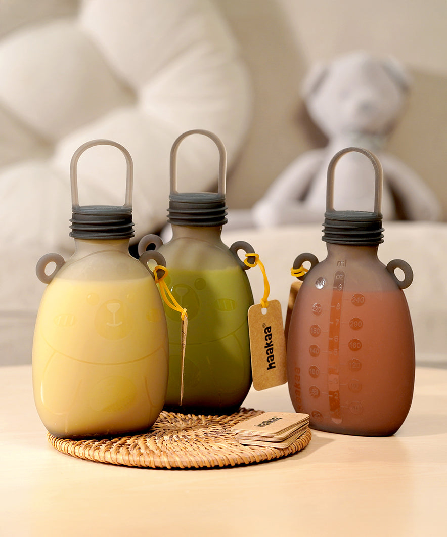 Three The Haakaa Happii Bear Silicone Goody Pouches on a table with a label attached to the bear ear on the bottle. The left pouch is filled with yellow liquid, the middle is green liquid, and the right is orange/red liquid