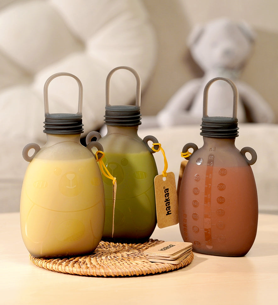 Three The Haakaa Happii Bear Silicone Goody Pouches on a table with a label attached to the bear ear on the bottle. The left pouch is filled with yellow liquid, the middle is green liquid, and the right is orange/red liquid