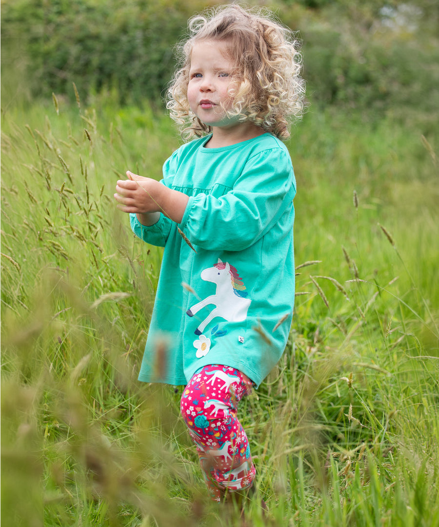 A child playing in a grassy field, wearing the Frugi Tilly Outfit - Pacific Aqua/Wild Horses