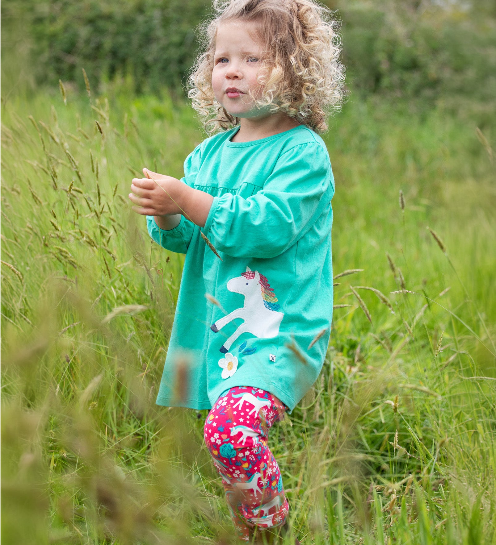 A child playing in a grassy field, wearing the Frugi Tilly Outfit - Pacific Aqua/Wild Horses