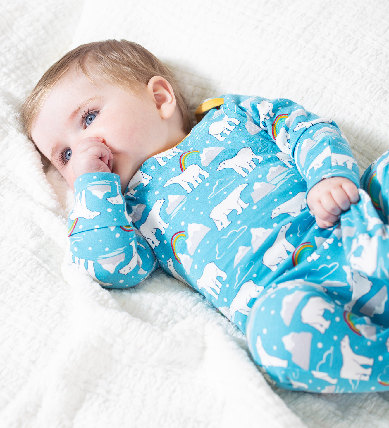 A baby lying on a white blanket, wearing the Frugi Easy Dressing Babygrow - Winter Tails