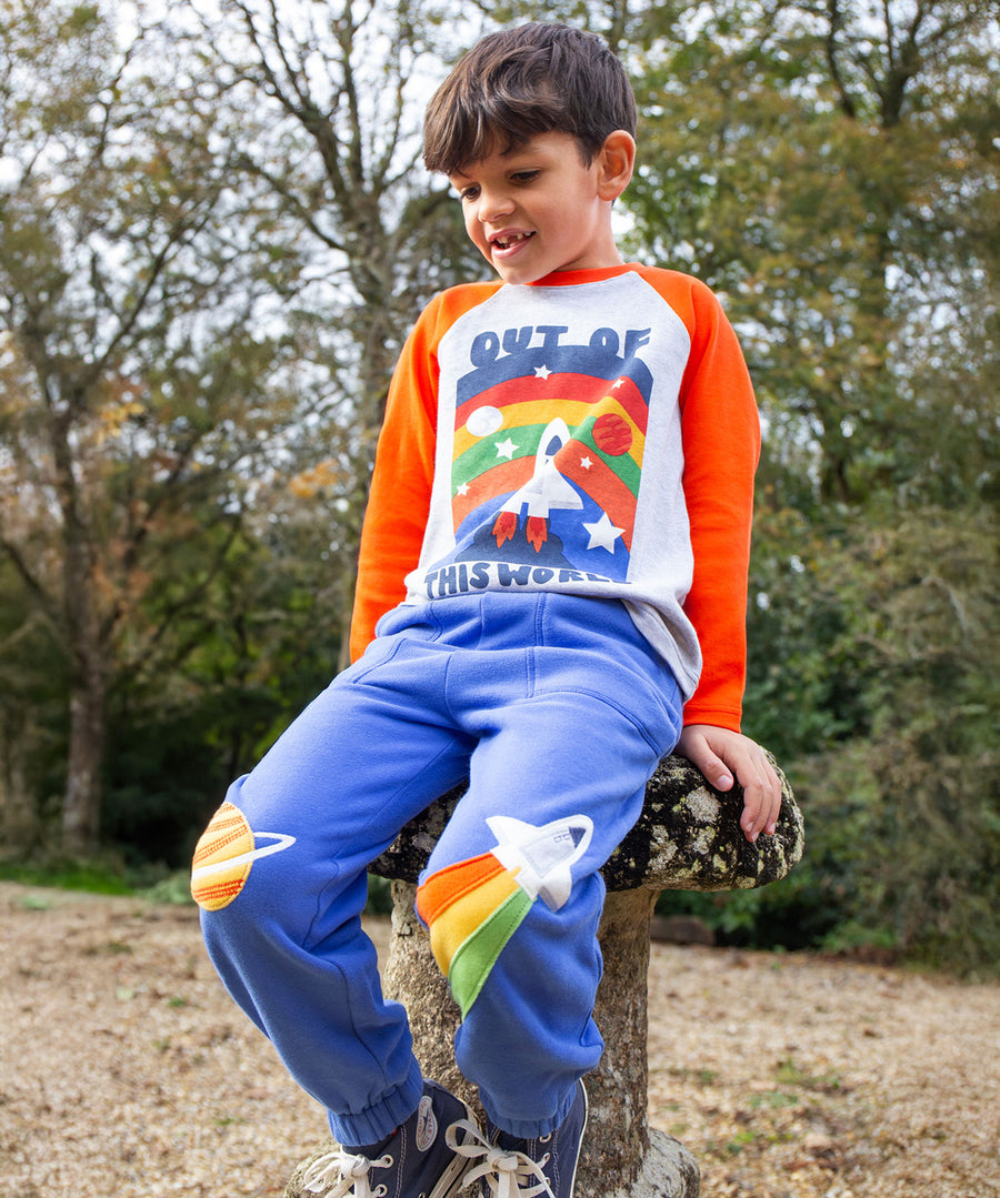 A child outside, sitting down on a stone stump, wearing the Frugi Malpas Applique Joggers - Blue Tang/Rocket and the Frugi Out Of This World top