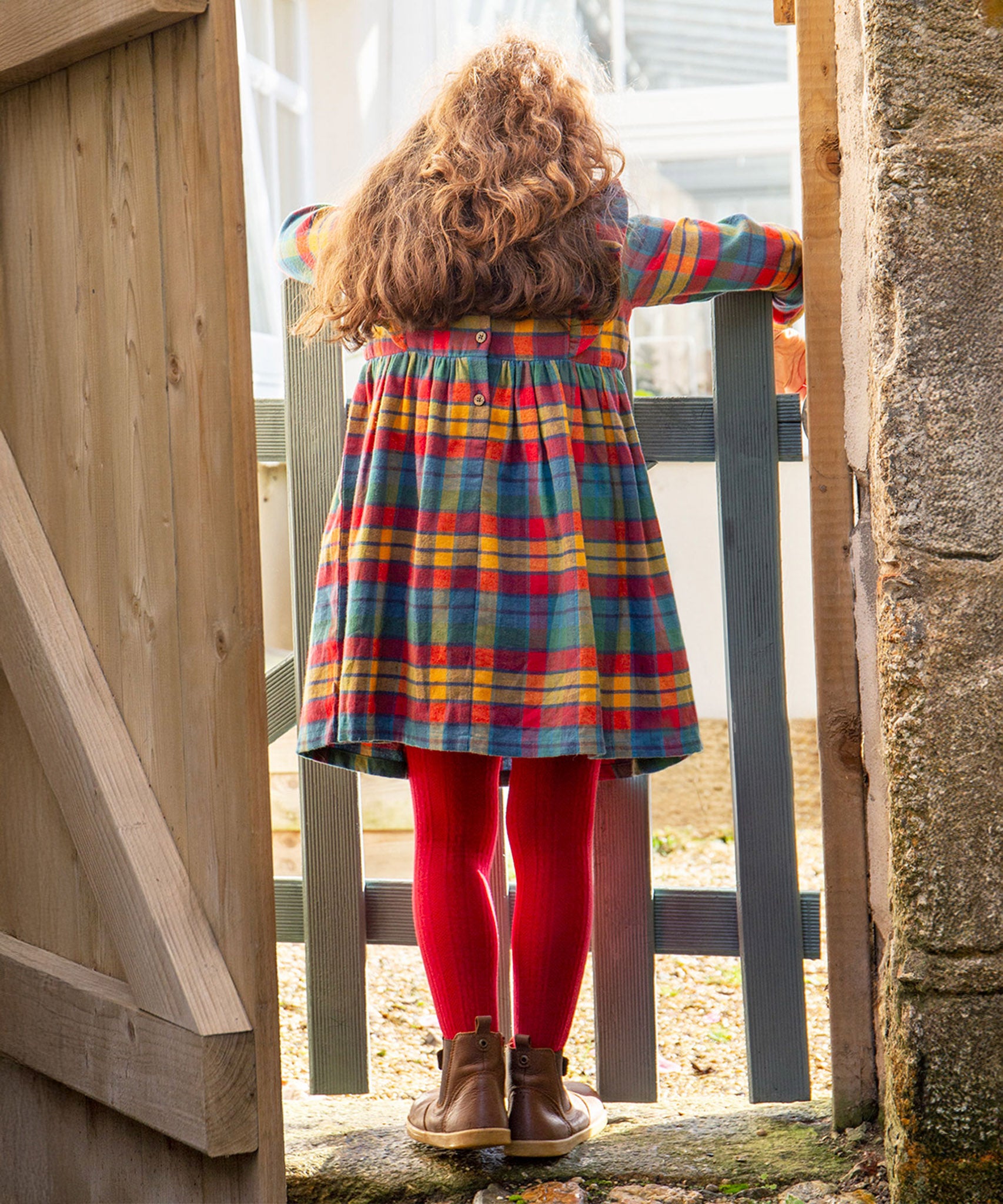 A child stood at a wooden gate, showing the back of the Frugi Agnes Dress - Merry Rainbow Check, with buttons at the back