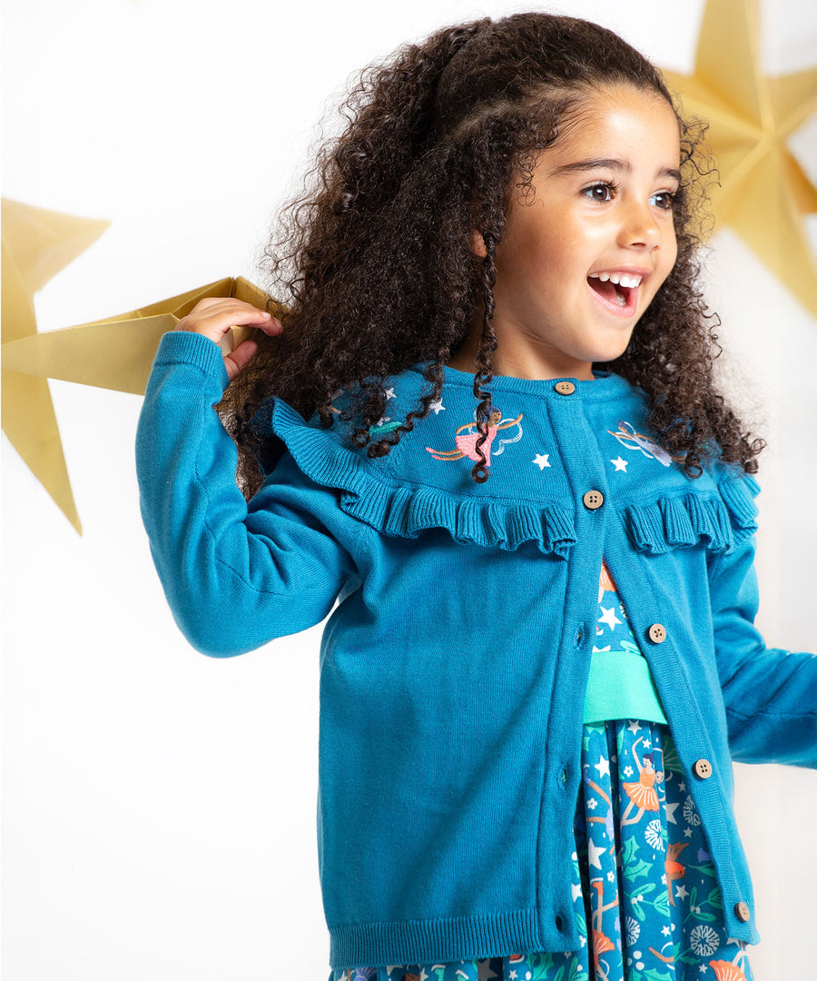 A happy child playing with gold, paper stars, wearing the Frugi Romi Ruffle Cardigan - Deep Water/Fairies. The cardigan is open showing a dress underneath