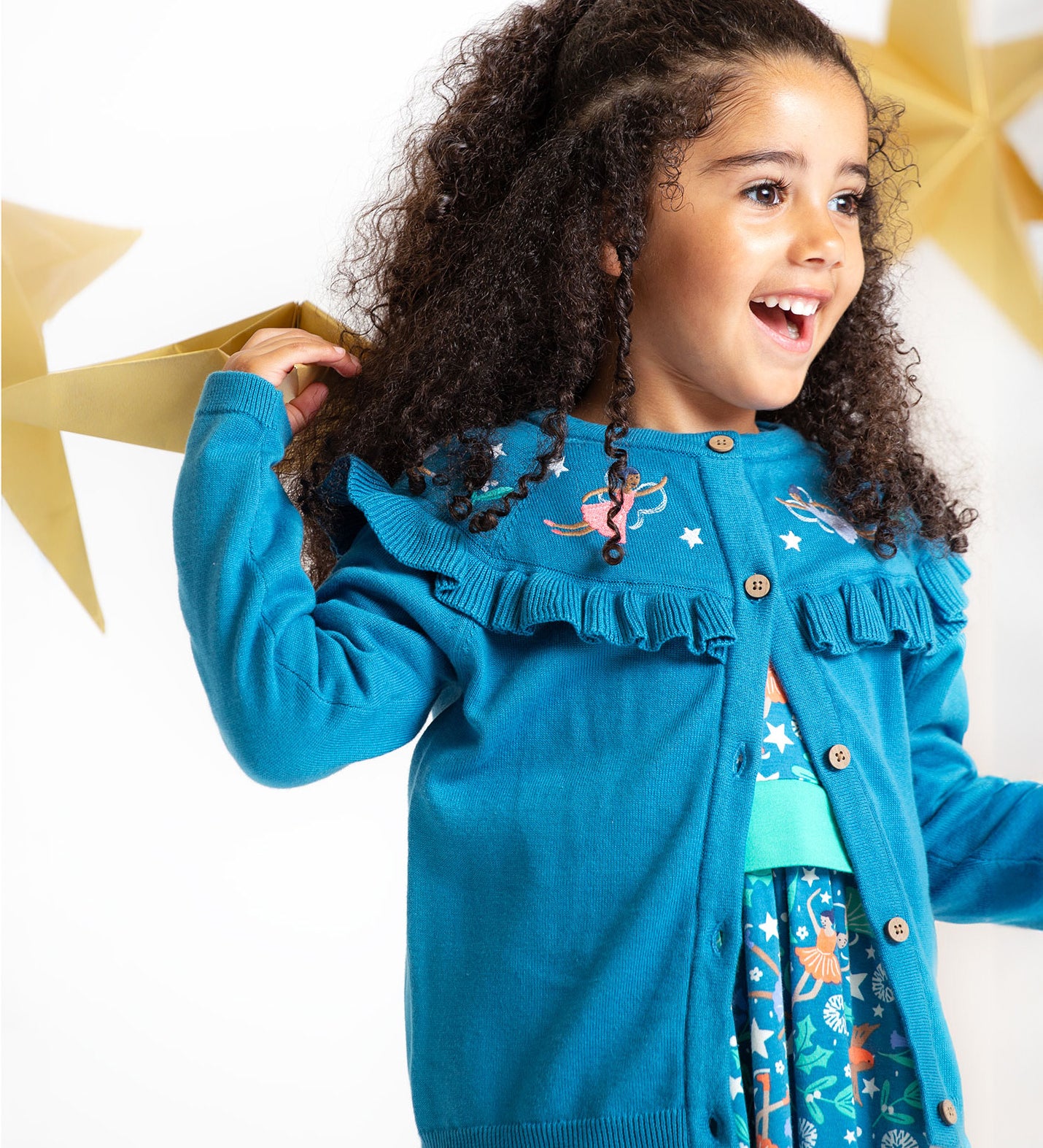 A happy child playing with gold, paper stars, wearing the Frugi Romi Ruffle Cardigan - Deep Water/Fairies. The cardigan is open showing a dress underneath