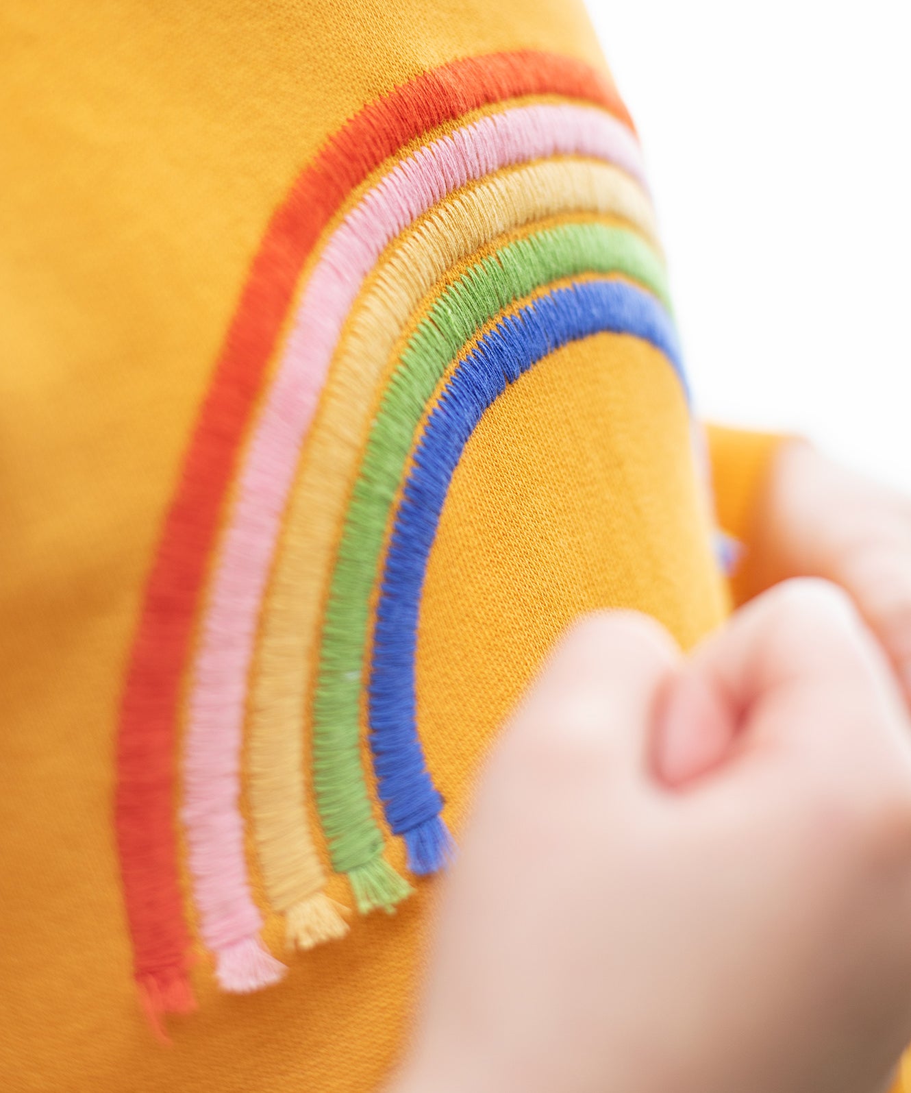 A closer look at the embroidery detailing and stitching of the Frugi Niamh Dress - Gold/Rainbow. The rainbow has a small, tassel at the bottom of each rainbow line