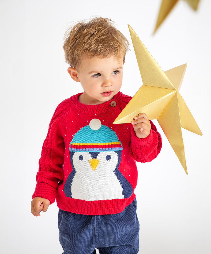 A child playing with a gold, paper star, wearing the Caleb Character Knit Jumper - True Red/Penguin. The jumper has a large penguin face and body, and the penguin is wearing a light blue hat