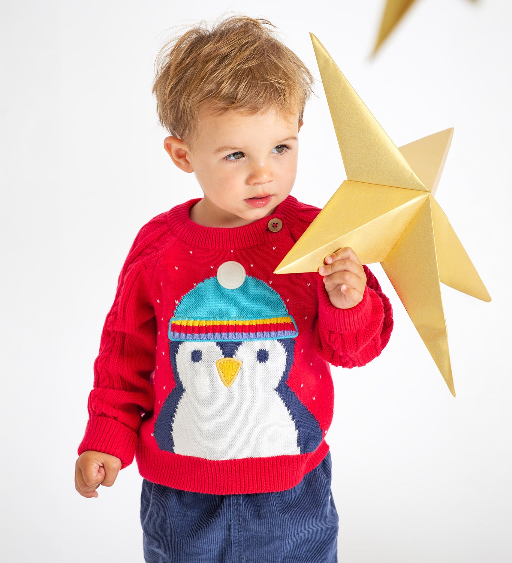 A child playing with a gold, paper star, wearing the Caleb Character Knit Jumper - True Red/Penguin. The jumper has a large penguin face and body, and the penguin is wearing a light blue hat