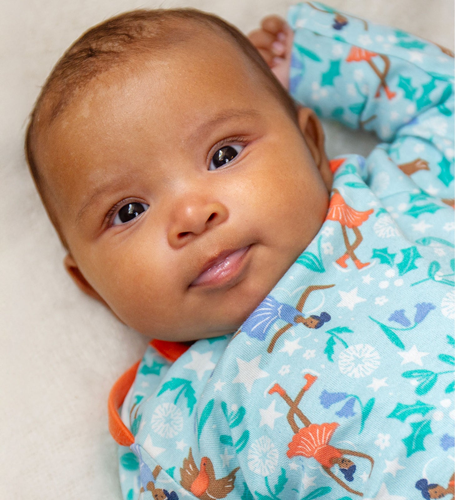 A baby lying down and looking forwards is wearing the Frugi Easy Dressing Printed Babygrow - Enchanted Forest. The image shows the ice skating fairies, robin and flower print on a blue fabric