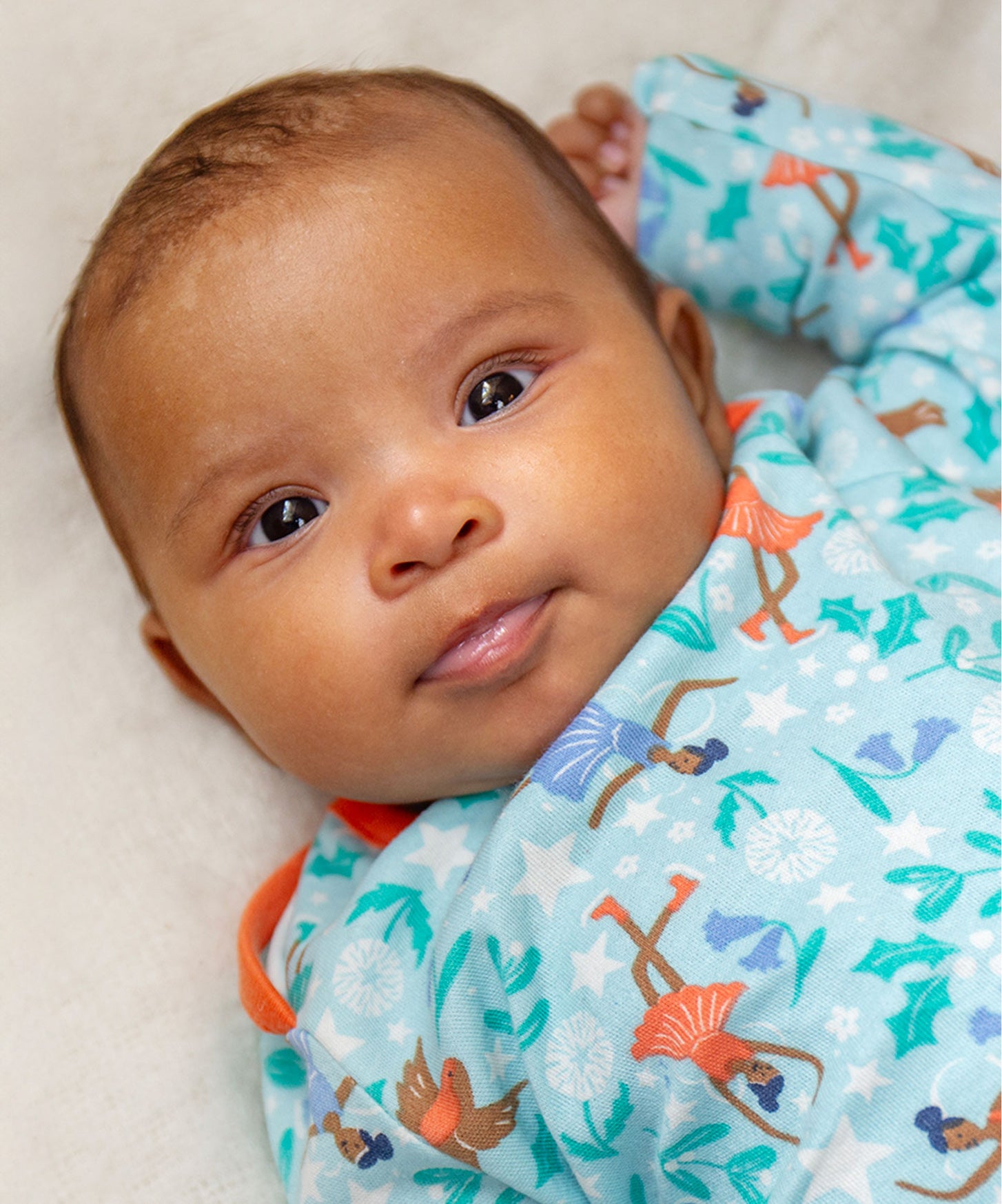 A baby lying down and looking forwards is wearing the Frugi Easy Dressing Printed Babygrow - Enchanted Forest. The image shows the ice skating fairies, robin and flower print on a blue fabric