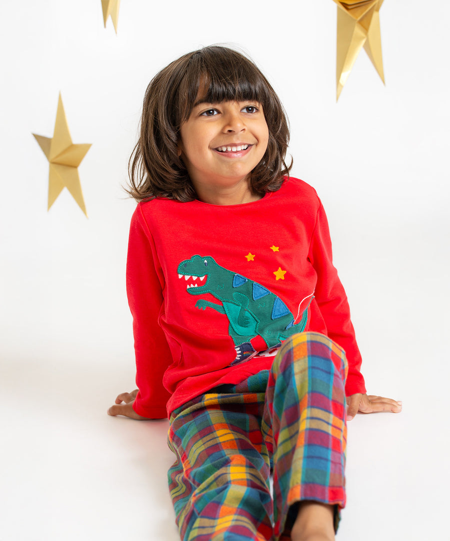 A child sat on the ground, smiling, and wearing the Frugi Caden Pyjamas - Merry Rainbow Check/Dinosaur with a white and gold star background