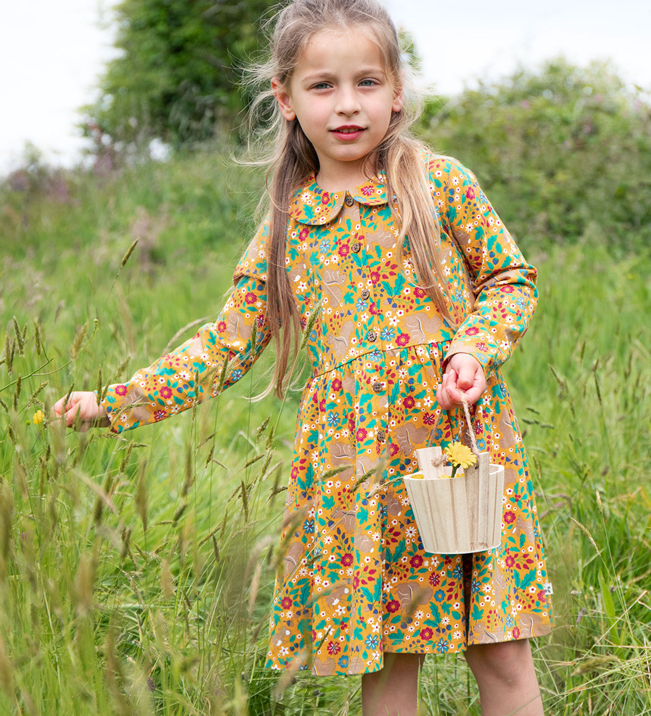 A child outside, holding a wooden basket and wearing the Frugi Autumn Dress - Autumn Friends