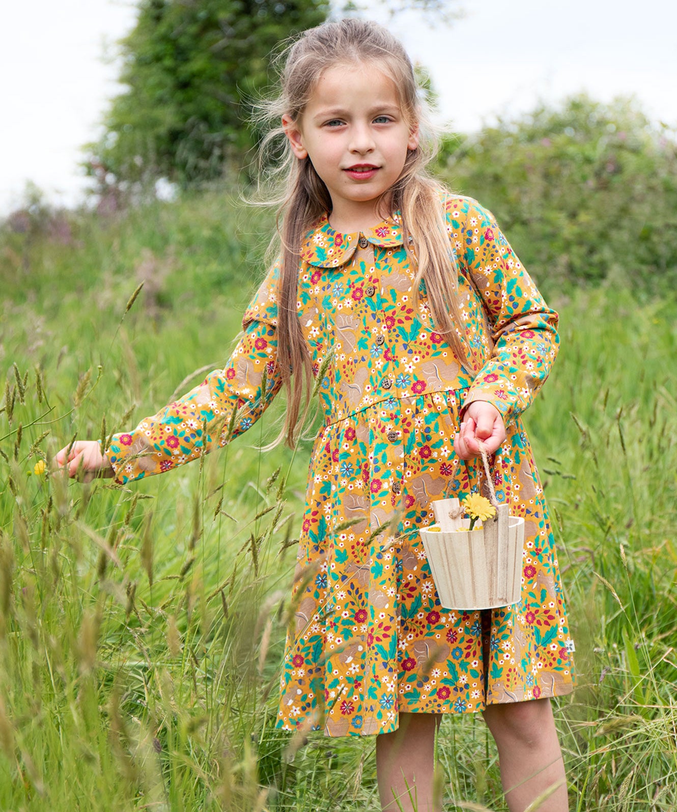 A child outside, holding a wooden basket and wearing the Frugi Autumn Dress - Autumn Friends