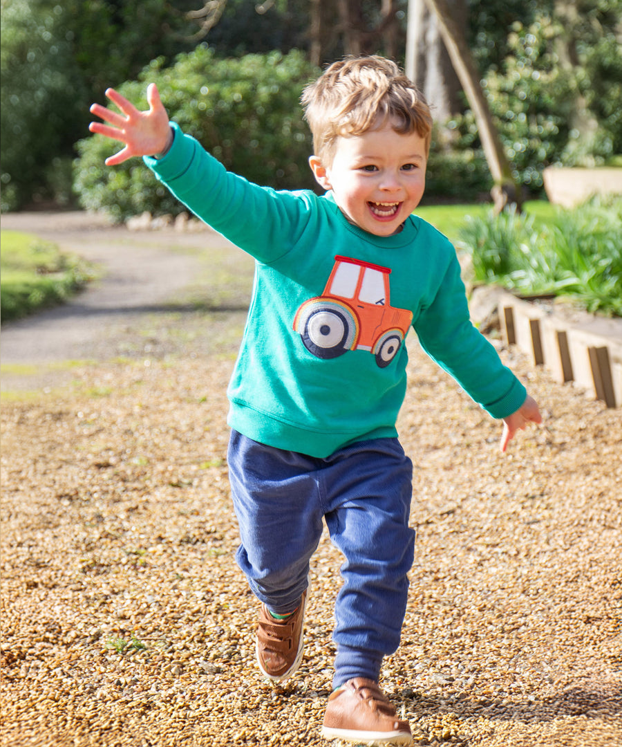 A child playing outside with their arms spread wide, wearing the Frugi Easy On Jumper - Iguana/Tractor