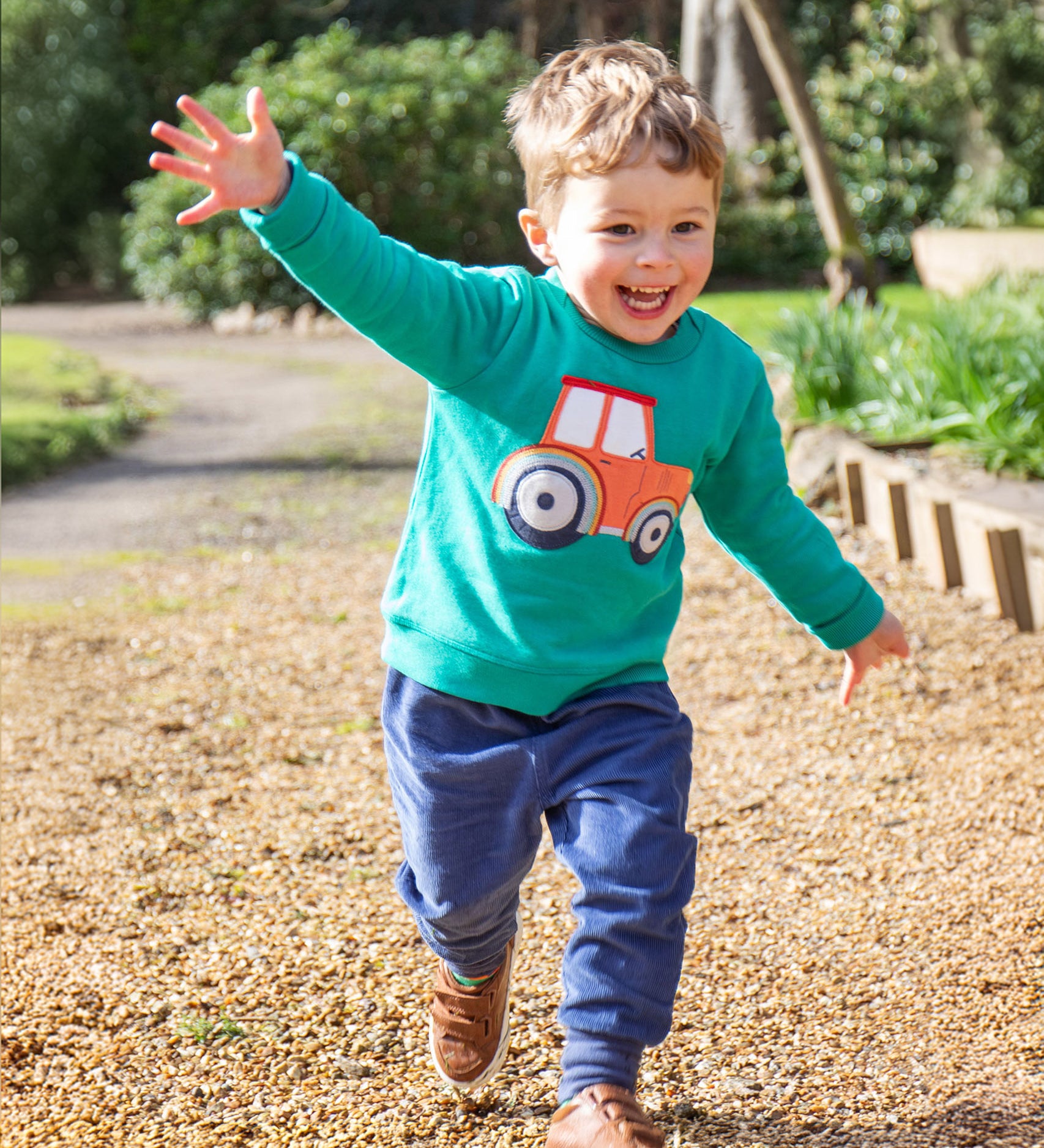 A child playing outside with their arms spread wide, wearing the Frugi Easy On Jumper - Iguana/Tractor