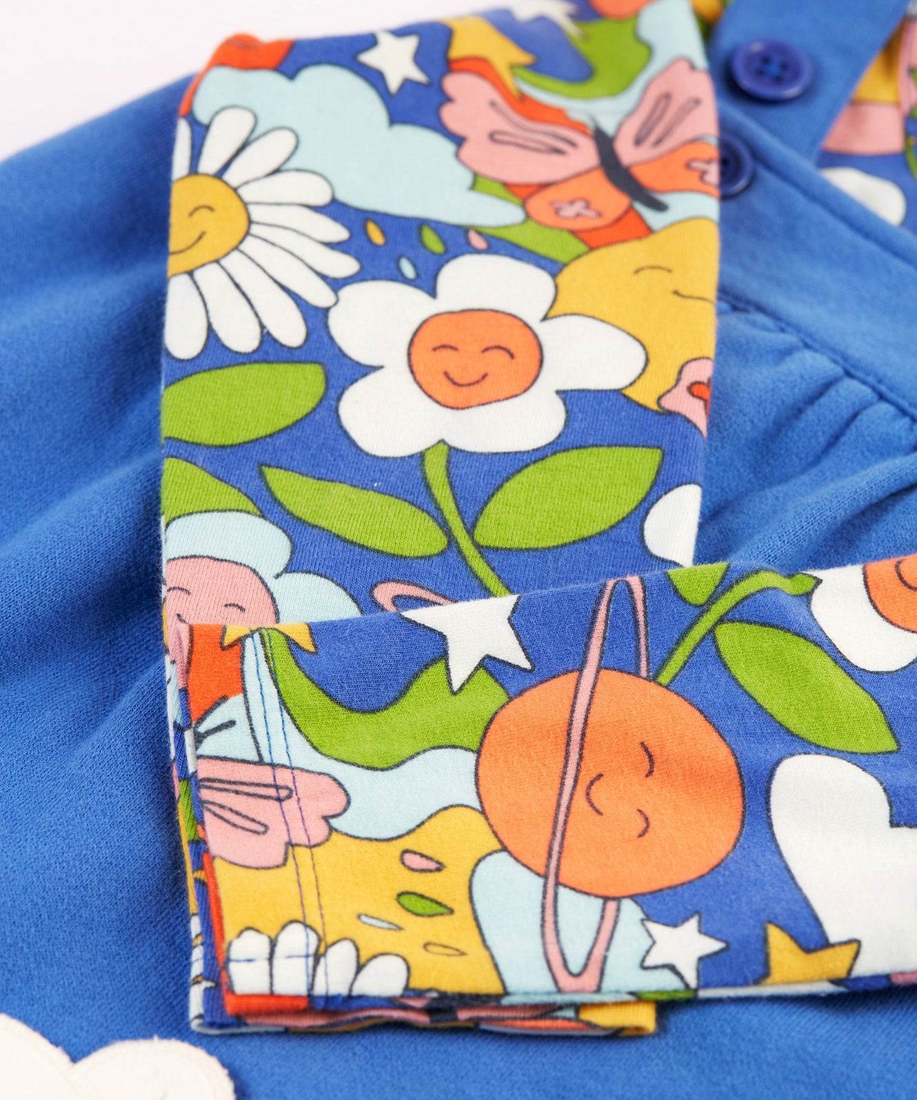 A closer look at the Retro Happy print on the long sleeve top of the Frugi Polly Pinafore Outfit - Blue Tang/Retro Happy. The Retro Happy print includes smiling planets, flowers, stars, clouds, bees and butterflies on a blue fabric which matches the dress