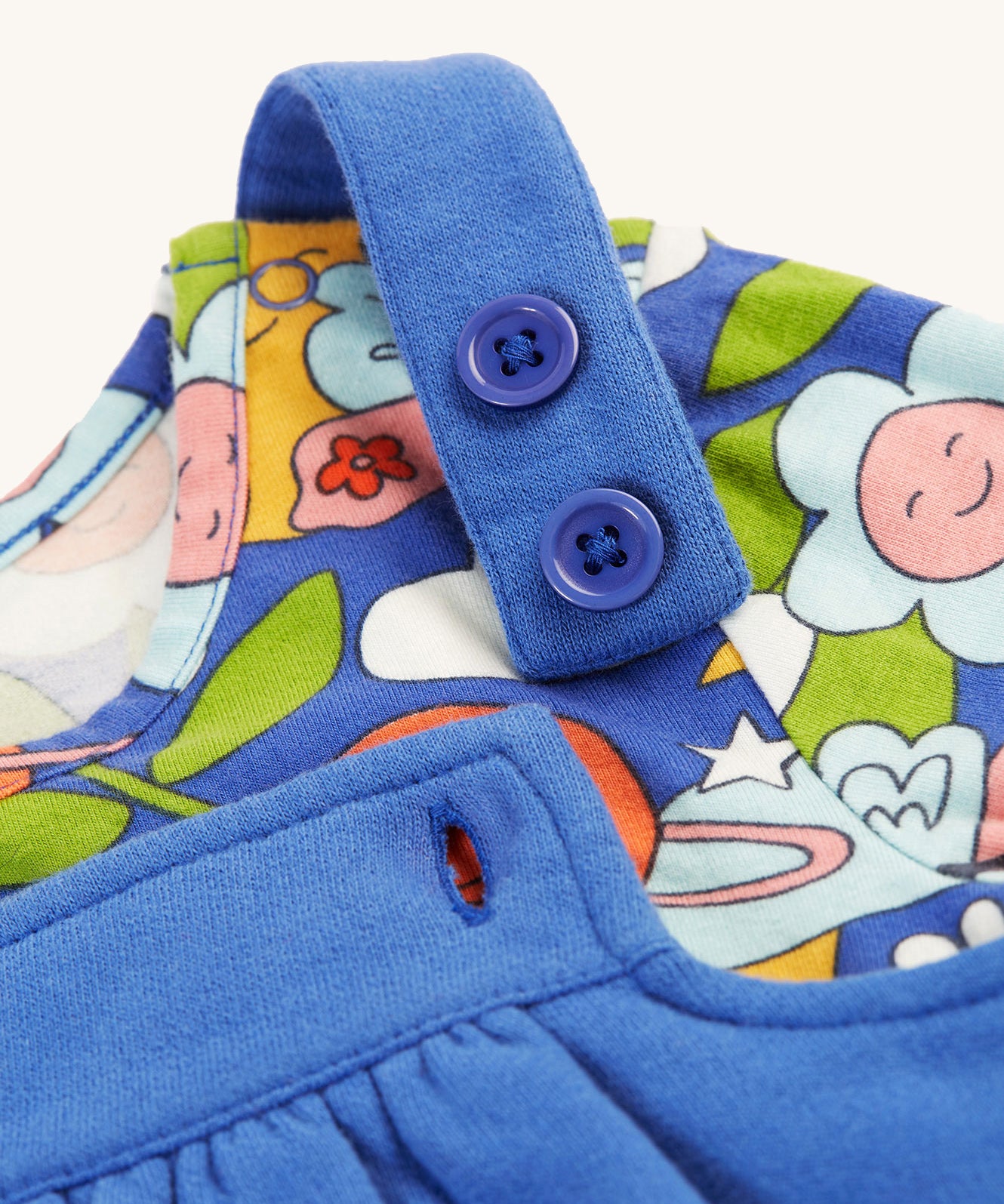 A closer look at the adjustable shoulder straps on the Frugi Polly Pinafore Outfit - Blue Tang/Retro Happy. The image also shows the Retro Happy print top, with its smiling planets, flower, cloud, bee and butterfly print.