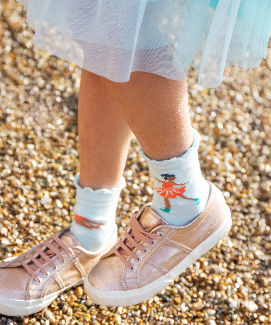 A child wearing pink trainers and a blue dress/skirt is wearing the light blue pair of Frugi Frill Socks 2 Pack- Enchanted Forest, with the ice skating fairy print