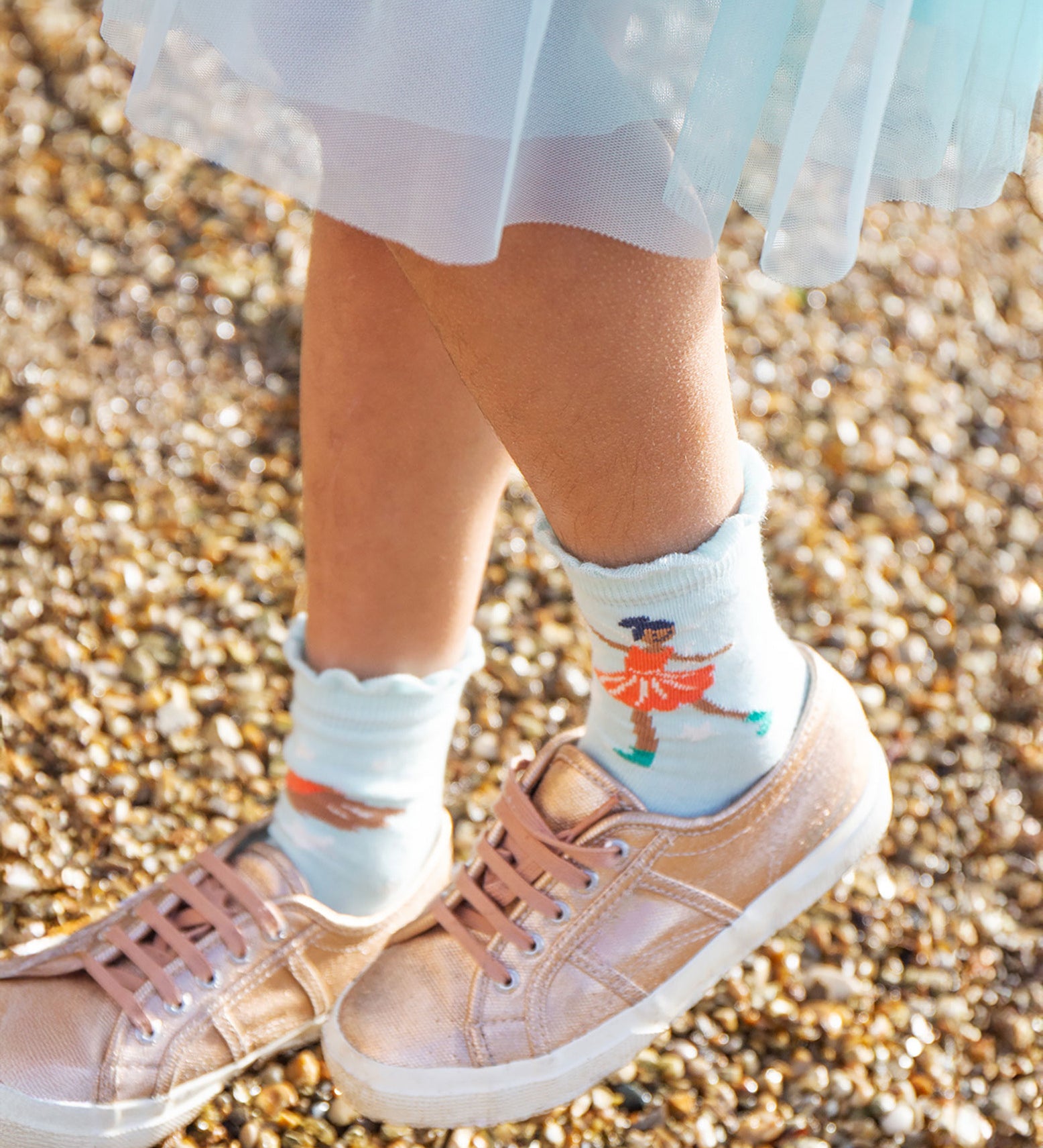 A child wearing pink trainers and a blue dress/skirt is wearing the light blue pair of Frugi Frill Socks 2 Pack- Enchanted Forest, with the ice skating fairy print