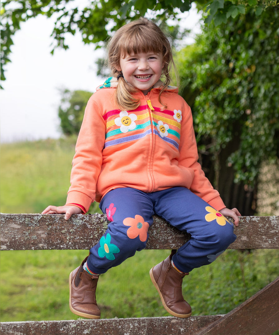 A child sat on a wooden fence, wearing the Frugi Perrin Parsnip Jogger - Navy Blue/Flowers, and a Frugi hoody