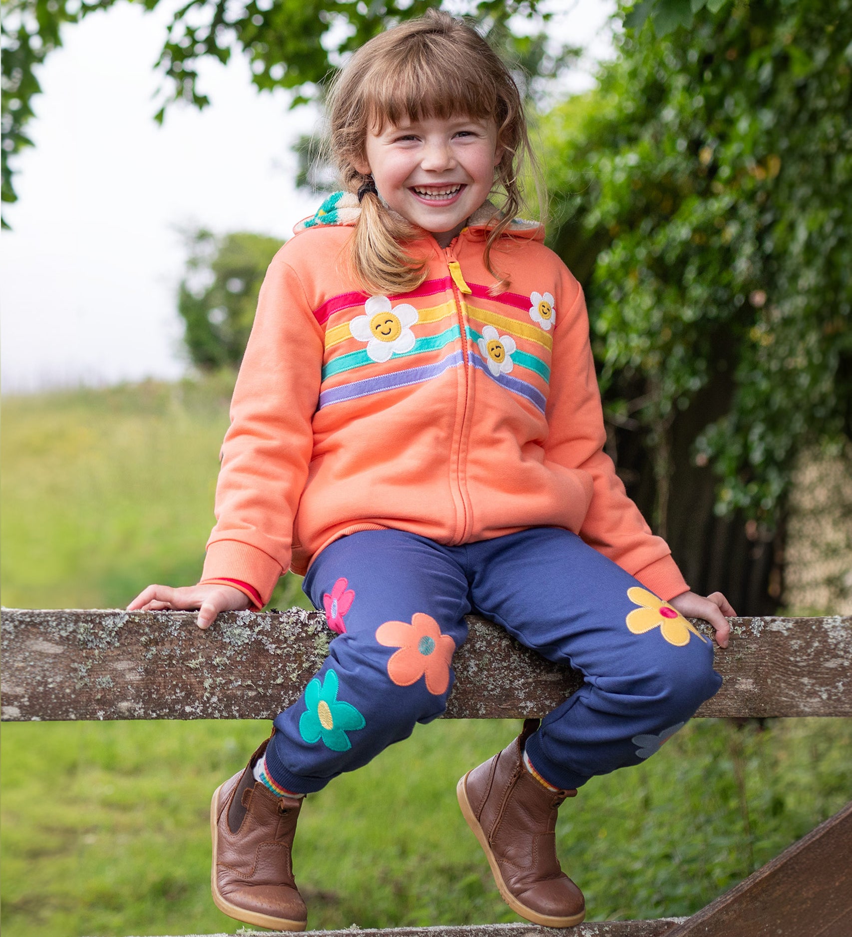 A child sat on a wooden fence, wearing the Frugi Perrin Parsnip Jogger - Navy Blue/Flowers, and a Frugi hoody