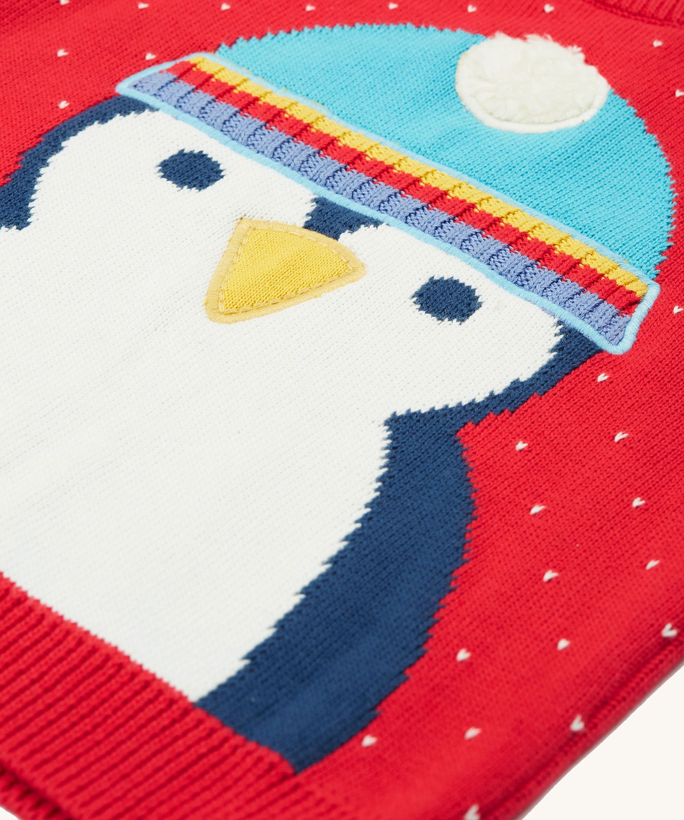 A closer look at the penguin character on the Caleb Character Knit Jumper - True Red/Penguin. The penguin has a navy blue and white body, yellow beak, and a light blue hat with white pom-pom on top 