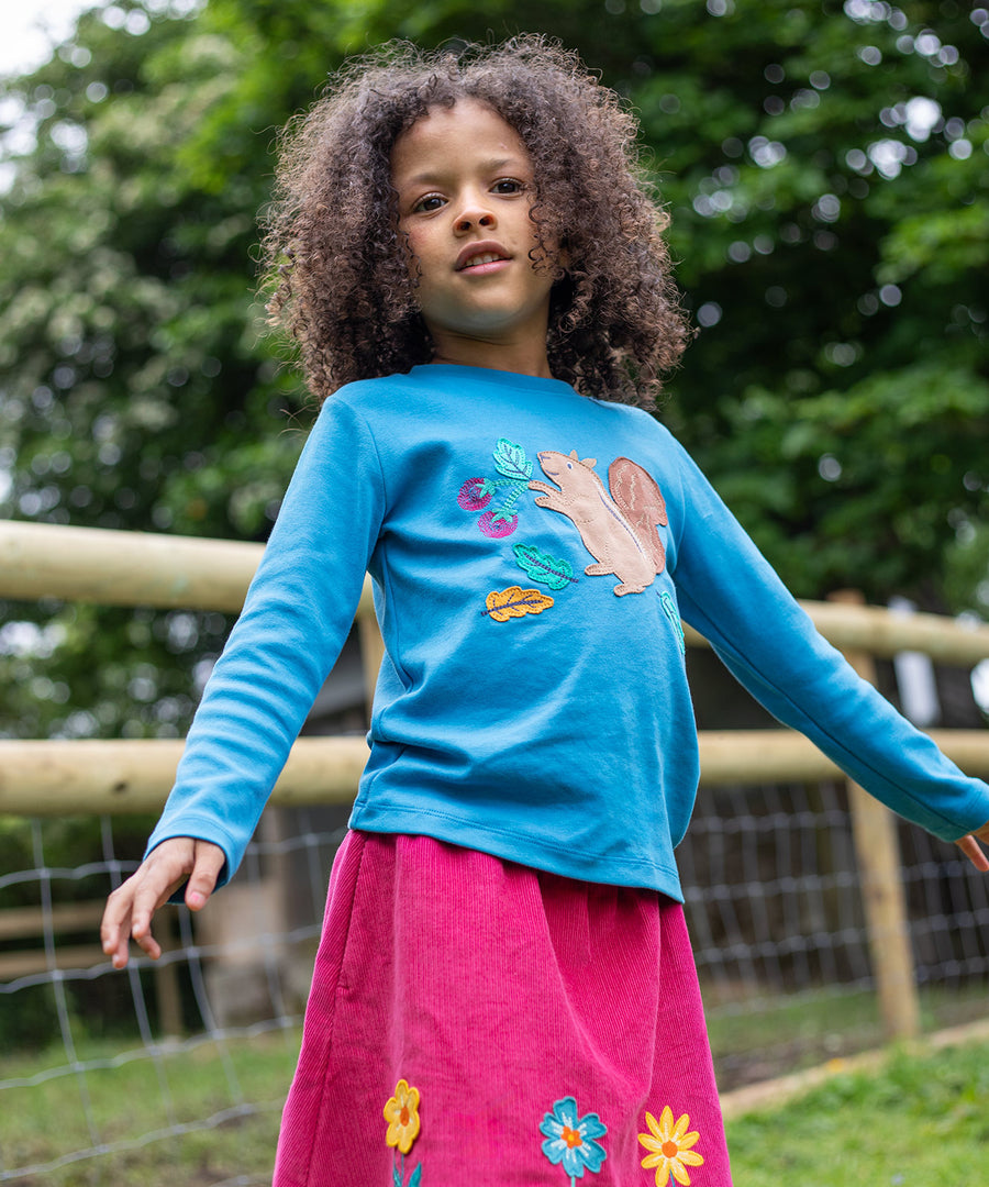 A child outside, wearing the Frugi Adventure Applique Top - Deep Water/Squirrel and a cord Frugi skirt