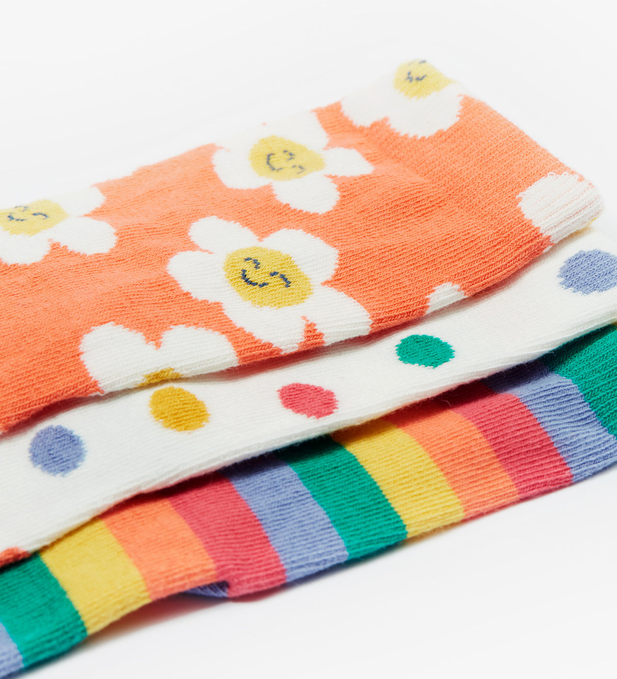 A closer look at the Frugi Rock My Socks - Stay Wild 3 Pack prints. The socks are laid on top of each other with the top sock being the orange and flower print, the middle sock is the white and polka dot print, and the bottom sock is the rainbow stripe print