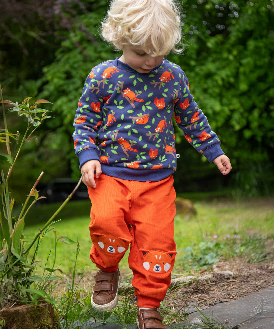 A child playing outside in a grassy field, wearing the Frugi Cassius Cord Trousers - Bonfire/Red Panda