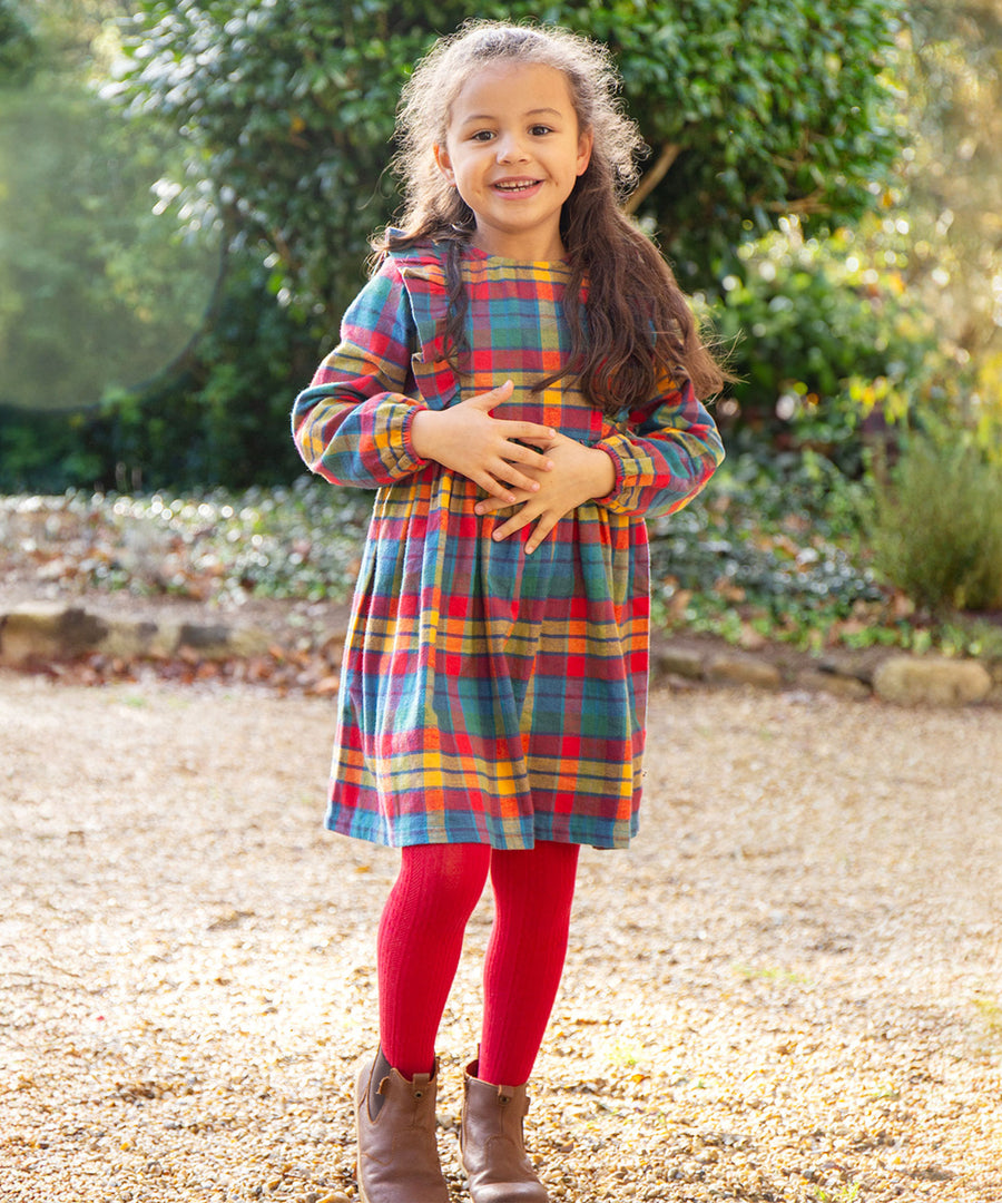 A child stood outside on a lawn with trees in the background, wearing the Frugi Agnes Dress - Merry Rainbow Check